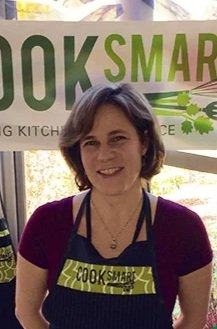Woman with short brown hair wearing a black apron and purple shirt, standing in front of a sign that reads 'COOK SMART'.