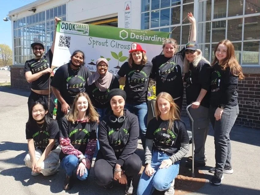 Group of people standing outside holding a banner for Sprout Circus, wearing matching black t-shirts with the circus logo, smiling at the camera.