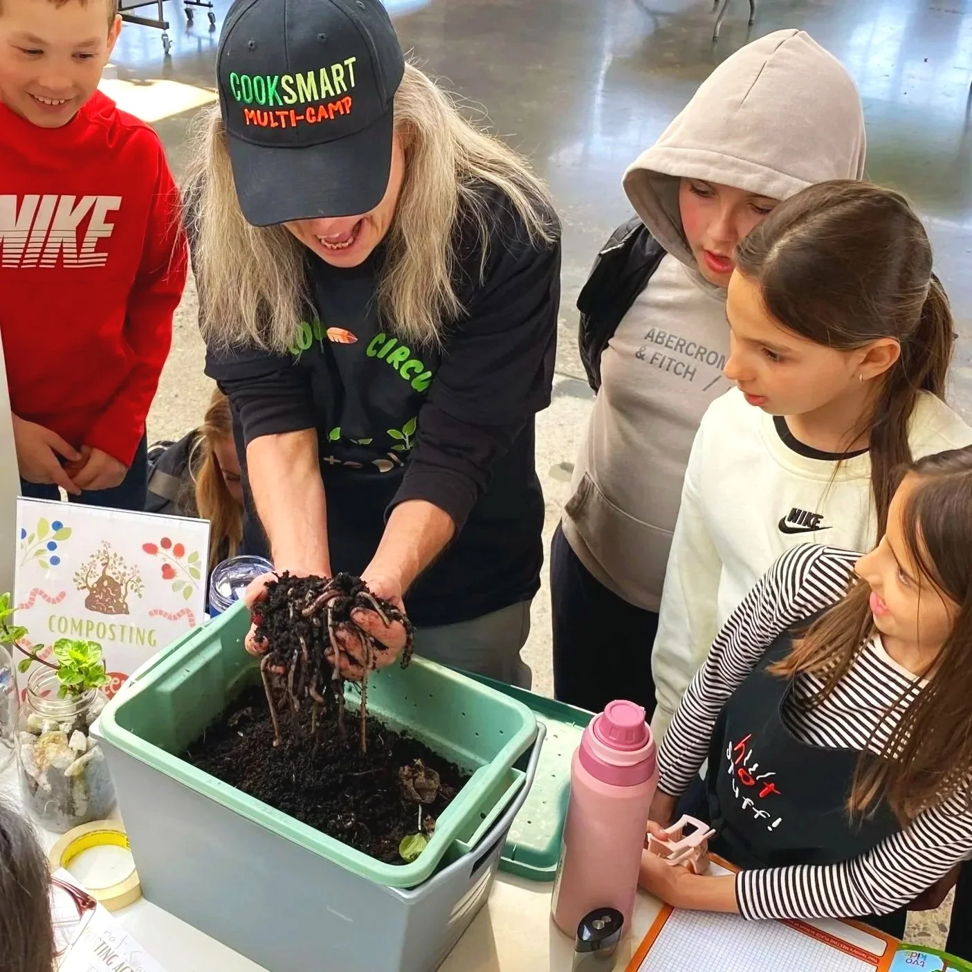 An adult woman showing children how to compost worms. The woman is holding a handful of dirt with worms, surrounded by children watching closely.
