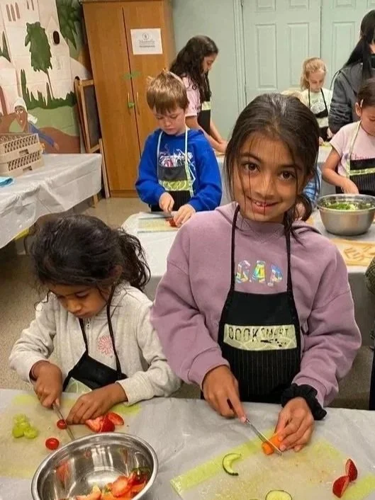 Children participating in a cooking class, chopping vegetables at a table in a classroom setting.