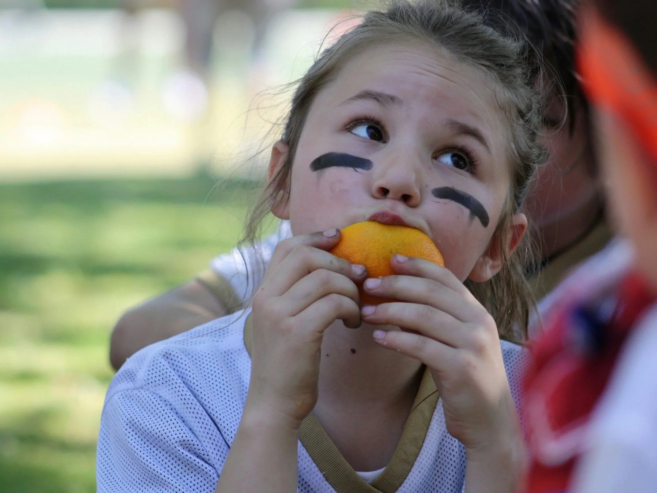 A young girl with face paint and black eye black is biting into an orange during an outdoor sports game or practice.