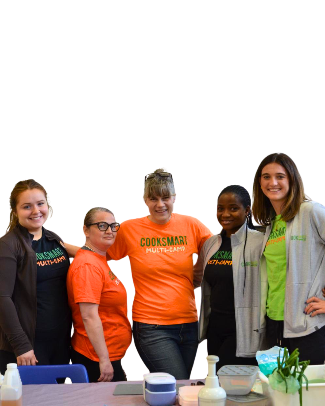 Group of five women standing together at a cooking event, wearing shirts that say CookSmart Multi-Camp, smiling. There are some cooking supplies and ingredients on the table in front of them.