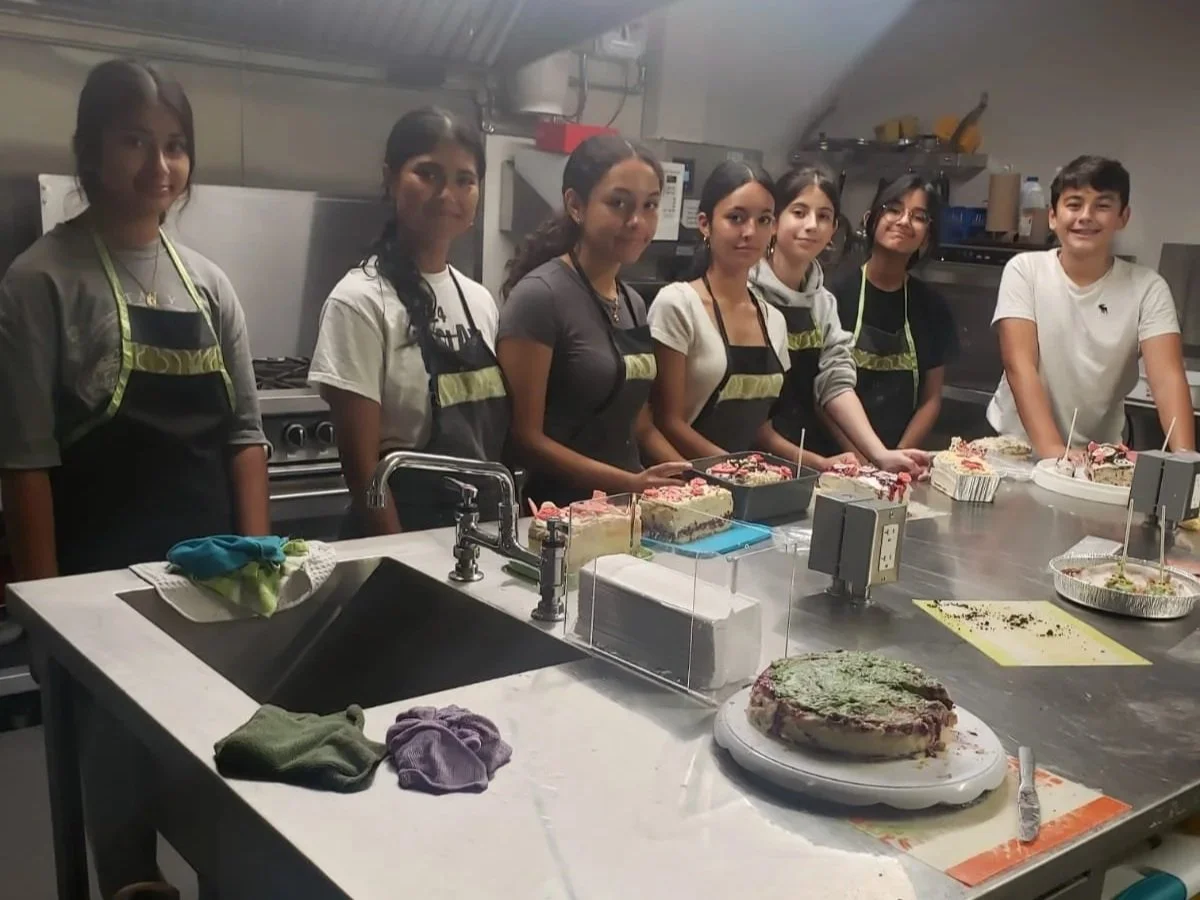 Group of young people in aprons standing behind a kitchen counter with various decorated cakes.
