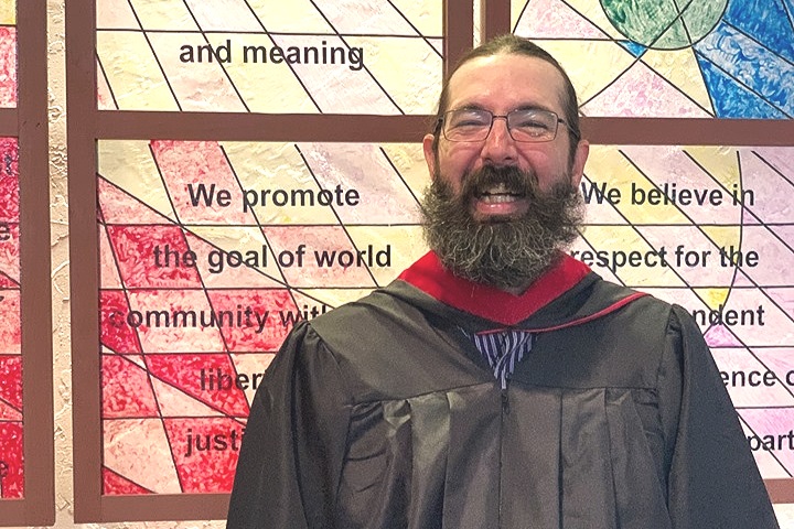 Rev. Andy Jacobs, on his ordination day, May 19th, 2019, stands before a stained glass created by Kirk Mackey depicting the UUA Seven Principles.