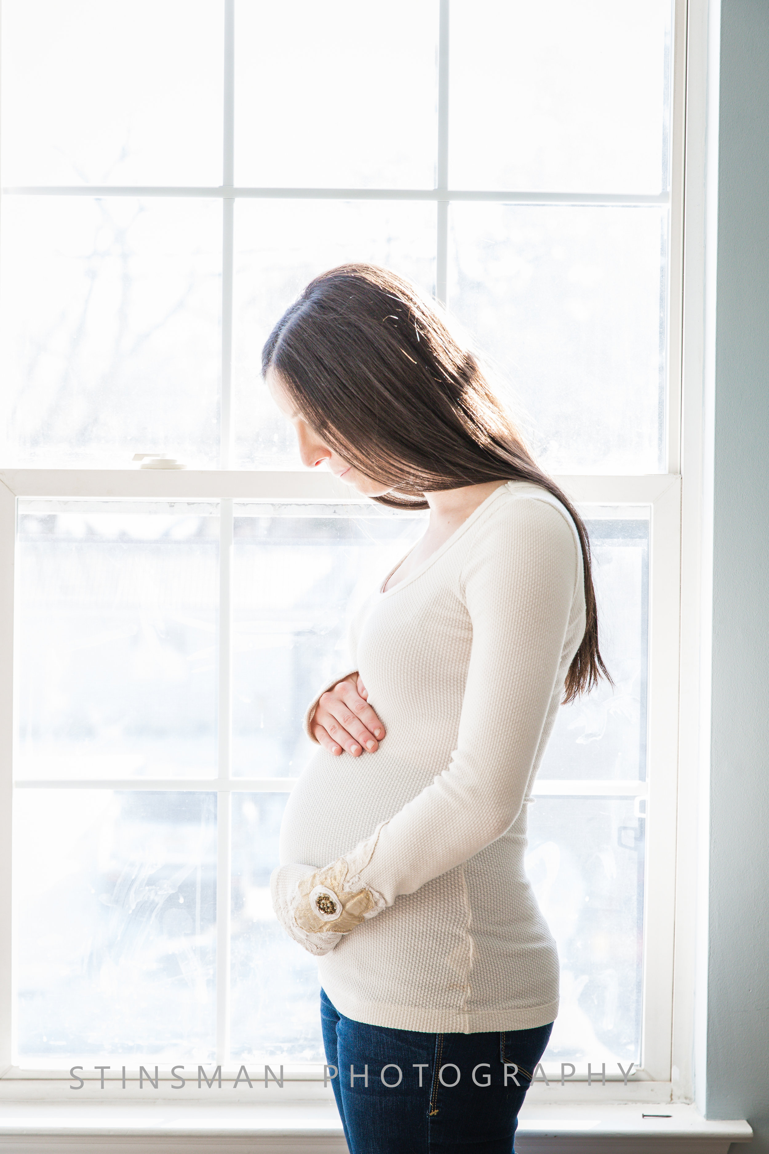 mom in front of a window cradling her pregnant belly wearing a white sweater