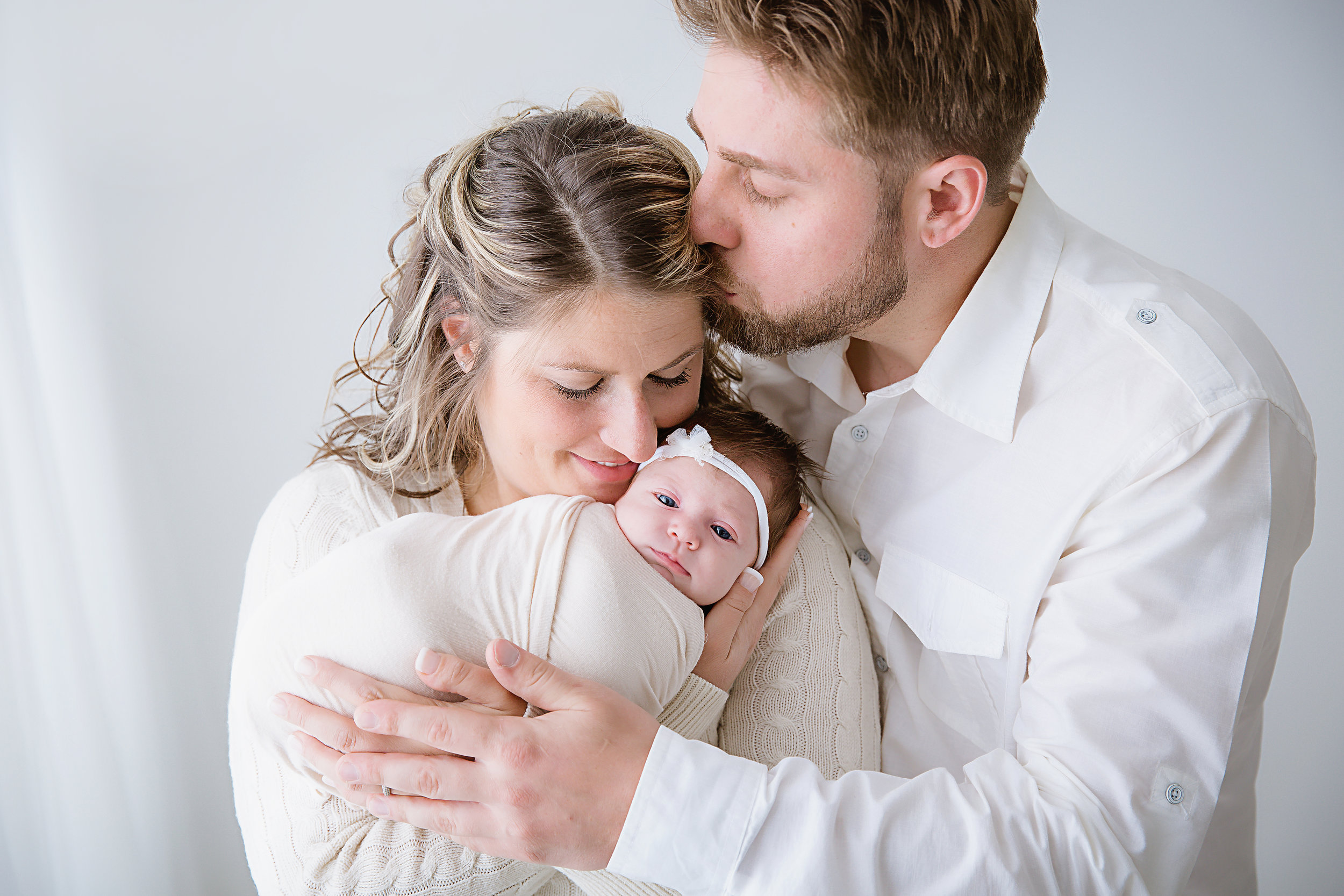new-parents-baby-girl-daughter-newborn-father-kissing-mommy-photography-shoot-white-studio-dinosaur-headband-new-jersey