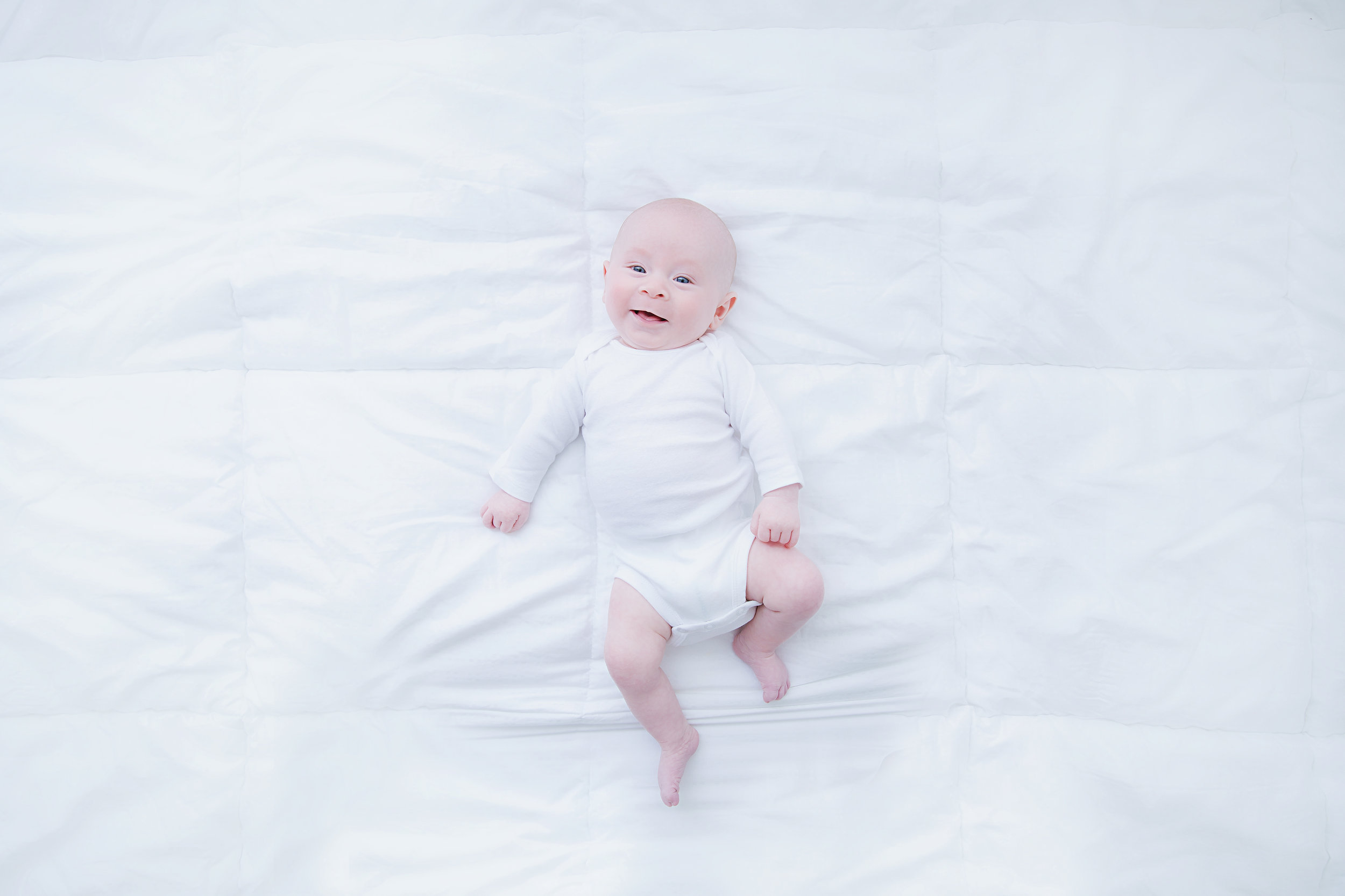 smiling-newborn-baby-lying-down-bed-birds-eye-photo-shoot-on-white-bed