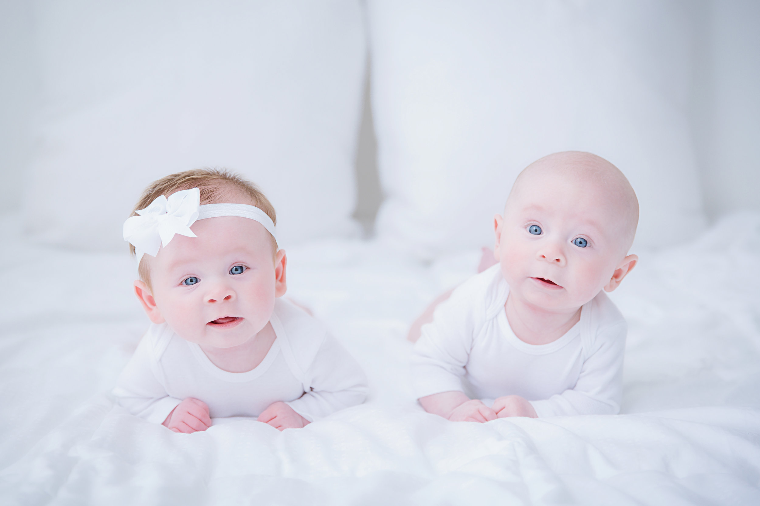 brother-sister-twins-3-months-old-baby-siblings-lying-on-tummy-looking-towards-camera-photography-shoot-new-jersey-white-bed-white-clothes