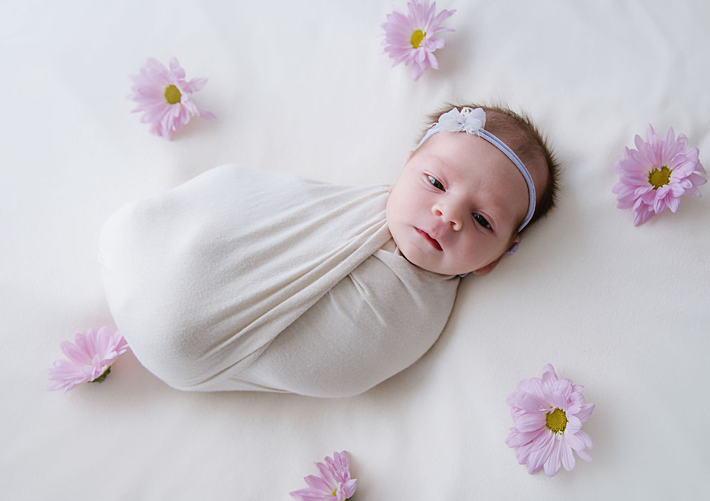 Purple flowers surrounding a wrapped up newborn baby girl. Both are lying on a soft surface in a photography studio in New Jersey.
