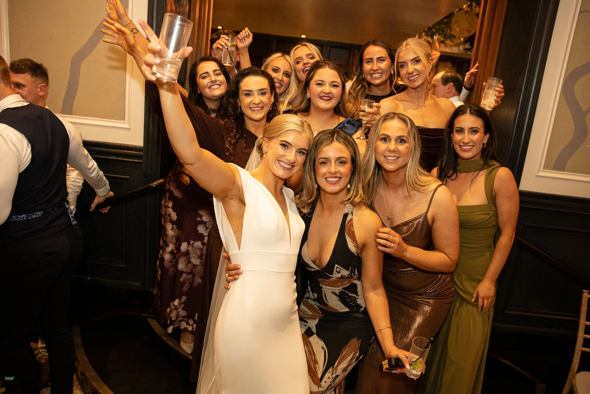 The Bride and her best friends gather during the evening drinks reception at Langtons Hotel, standing close together in warm, low lighting with glasses in hand. The bride, Kate Tobin, is at the centre in her white dress, smiling as the group poses fo