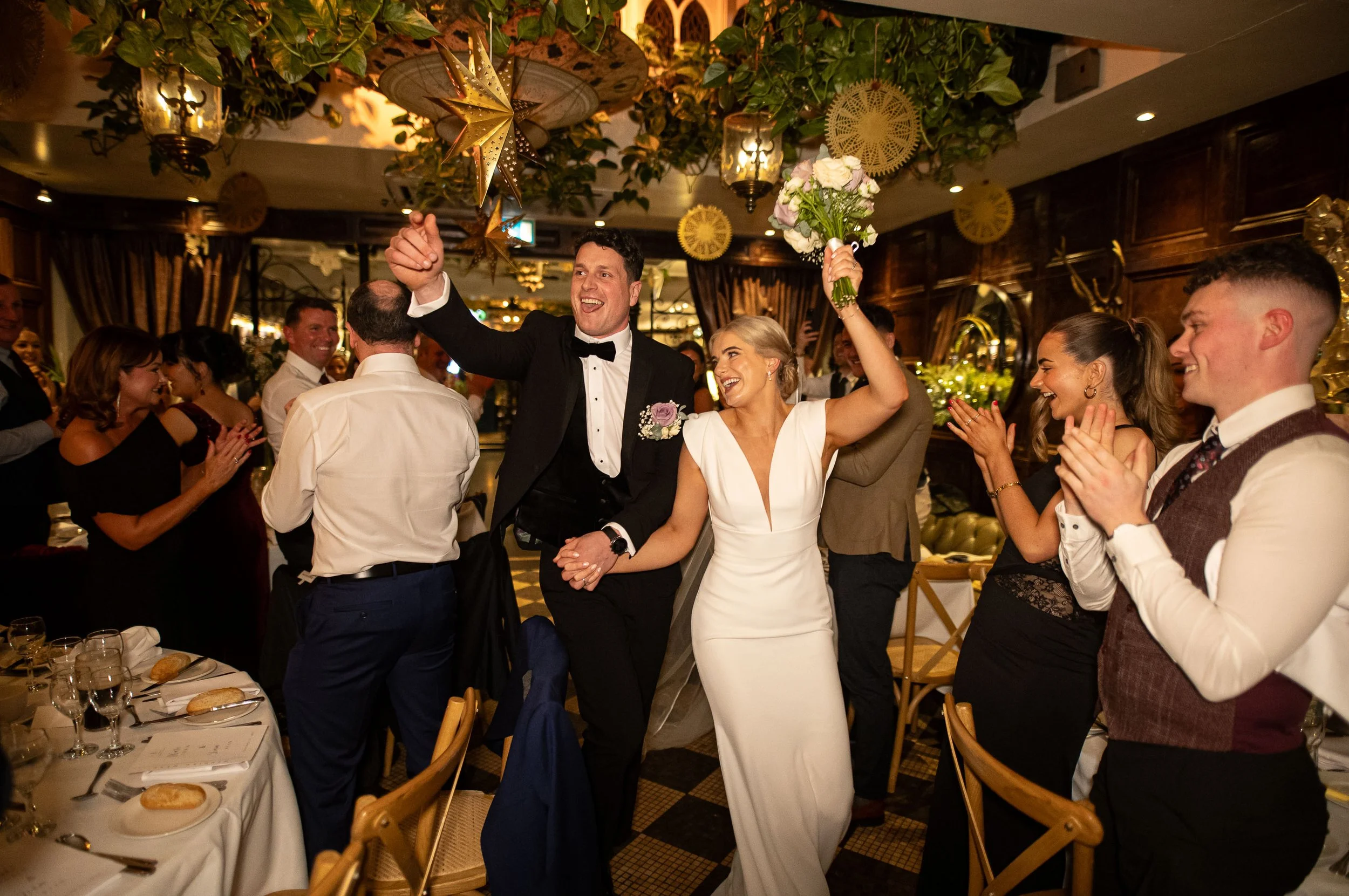 Newlyweds make their entrance into the ballroom at Langtons Hotel, walking hand in hand through a warm, golden-lit space filled with guests cheering and applauding. The bride lifts her bouquet in celebration while the groom smiles broadly. Members of
