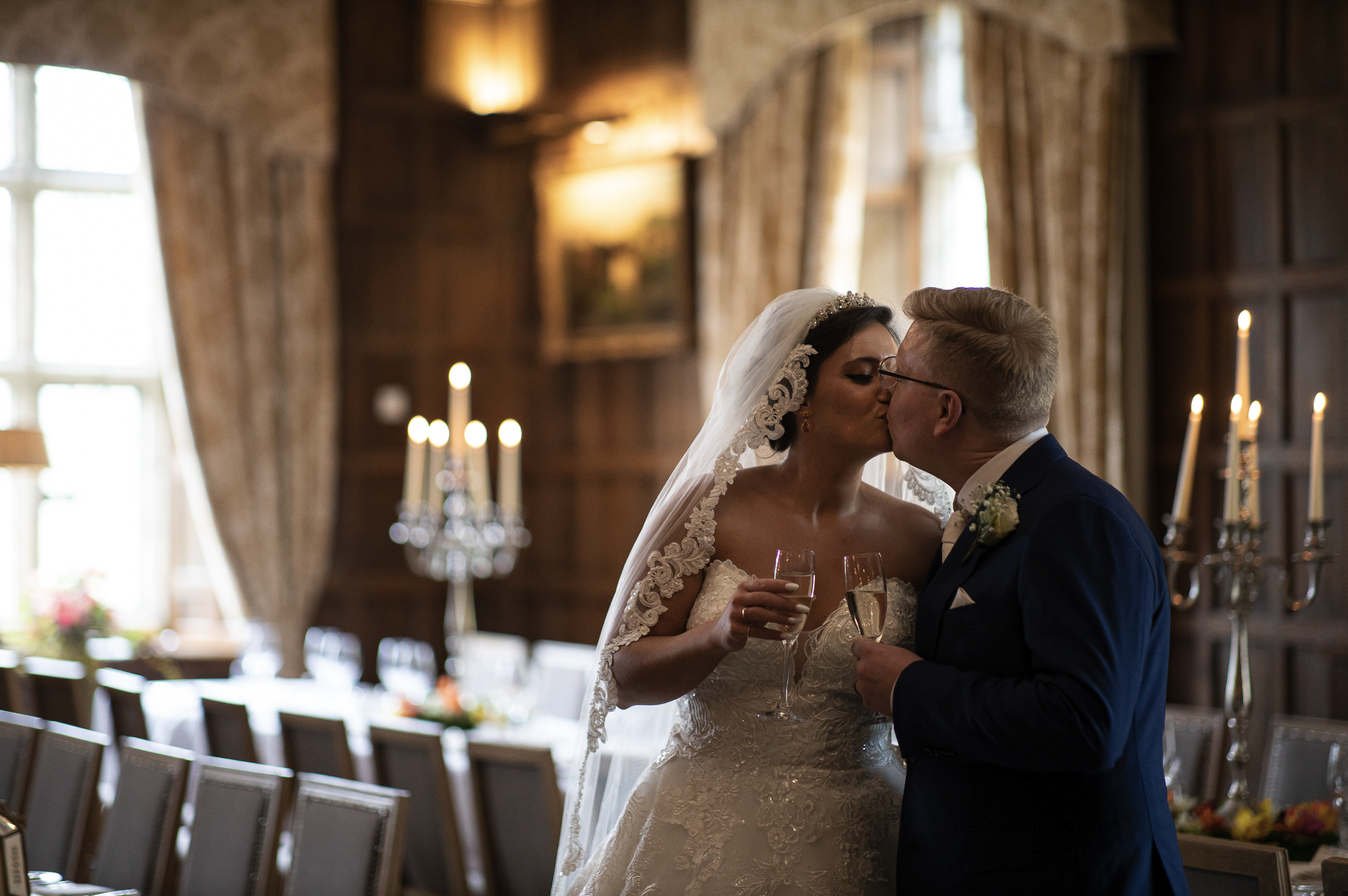 Bride and groom kissing indoors, holding champagne glasses, formal elegant setting.