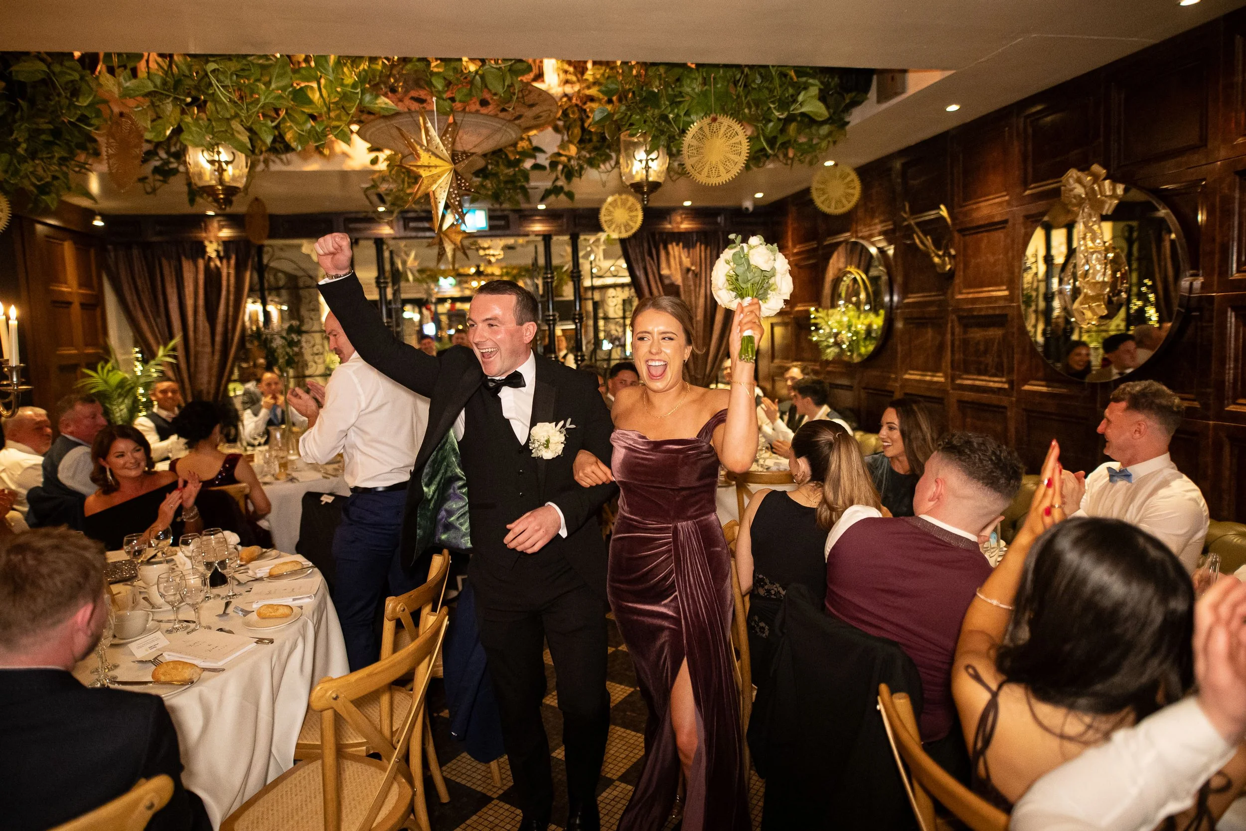 Bridesmaid and grooms man make their grand entrance into the ballroom at Langtons Hotel, walking hand in hand through a warm, golden-lit space filled with guests cheering and applauding. The bride lifts her bouquet in celebration while the groom smil