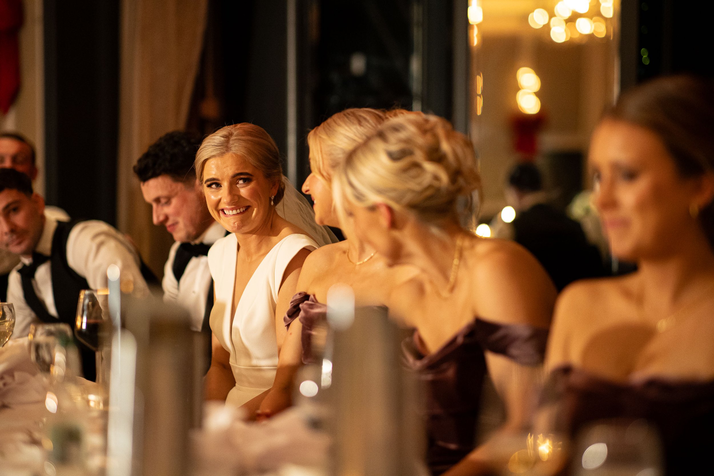 The groom stands at the head table in the Langtons Hotel ballroom, delivering his speech with a microphone in hand while the newlyweds and wedding party sit beside him, smiling and reacting warmly. Here we see the bride take a gasp while looking lovi