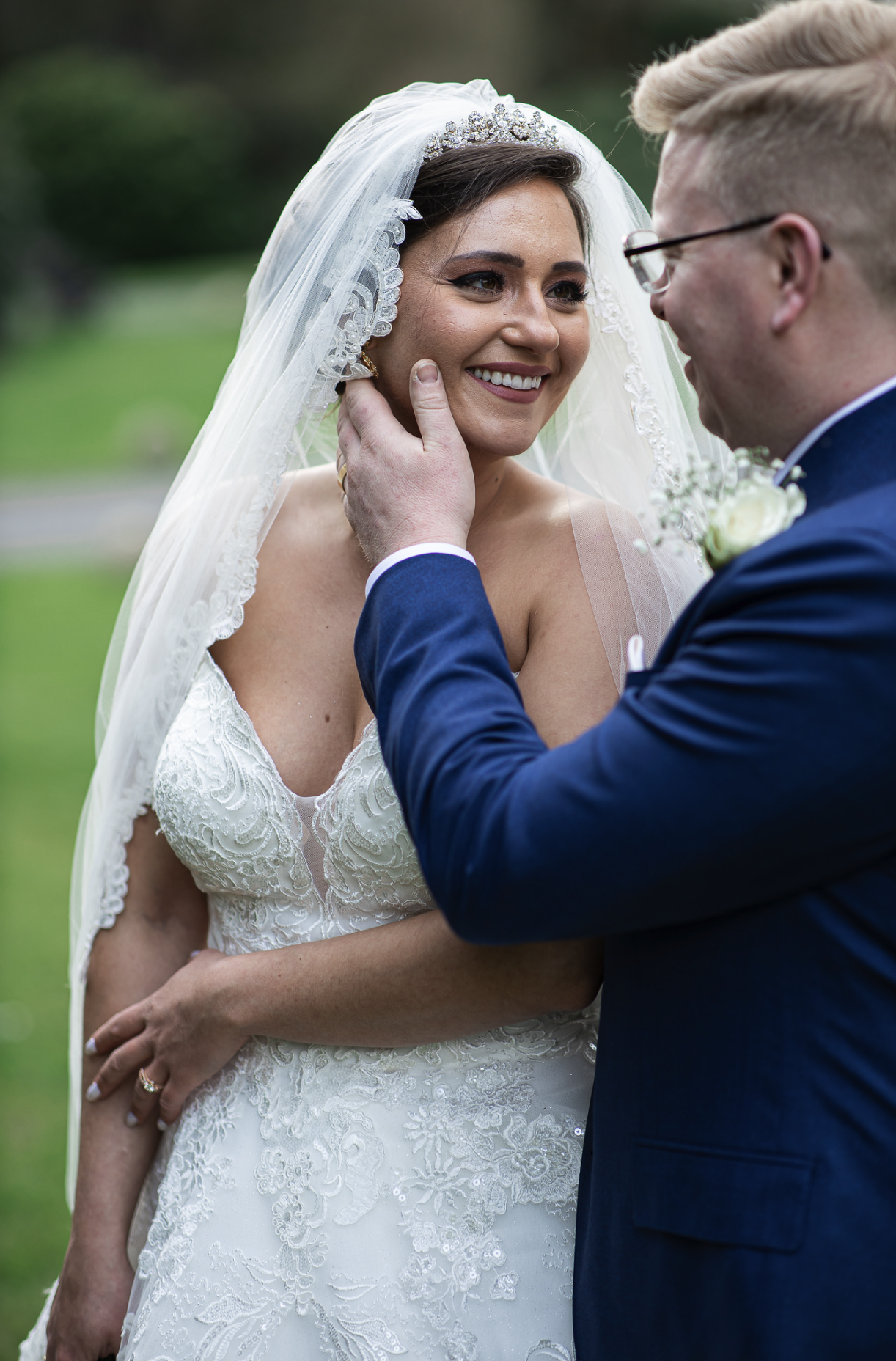Bride and groom in a loving embrace during a wedding outdoors, bride wearing a lace wedding gown and veil, groom in a blue suit.