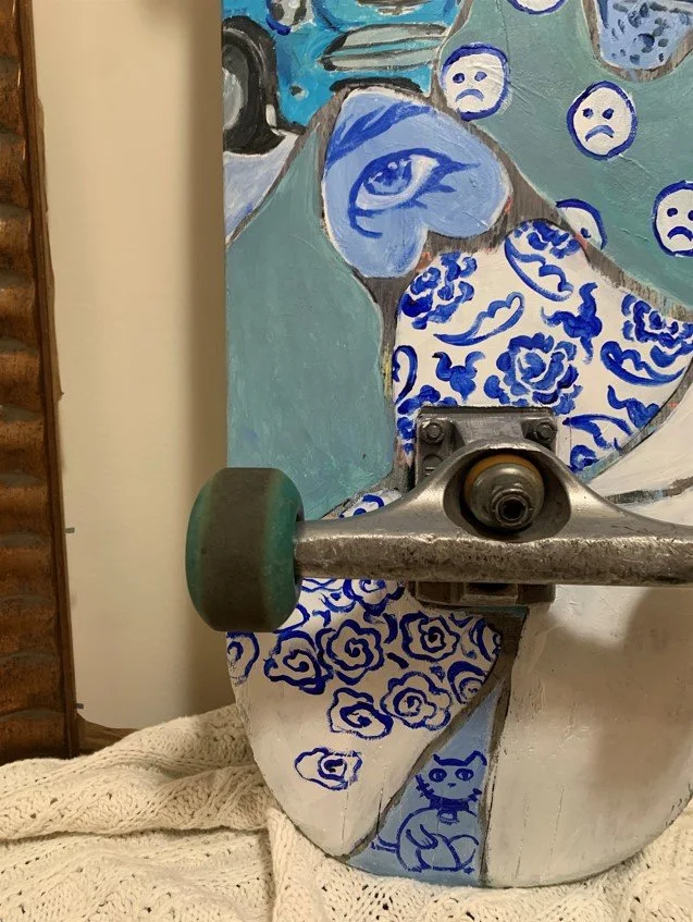 Image Description: Close up of flowers, eyes, faces, and cat painted in shades of blue on bottom of skateboard.