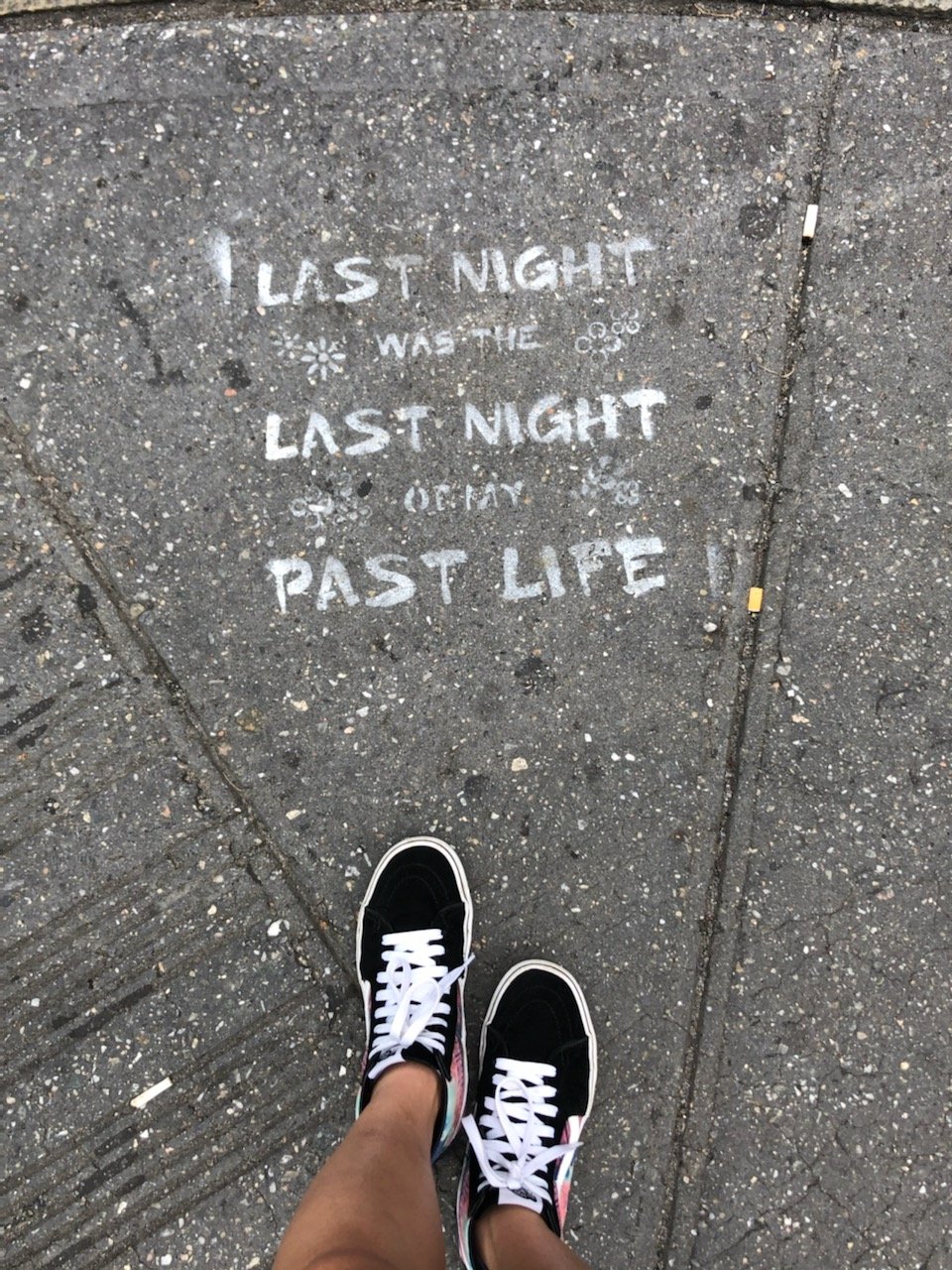 A picture of a grey concrete sidewalk. A pair of black and white sneakers stand in front of a spray painted sign that reads "last night was the last night of my past life".