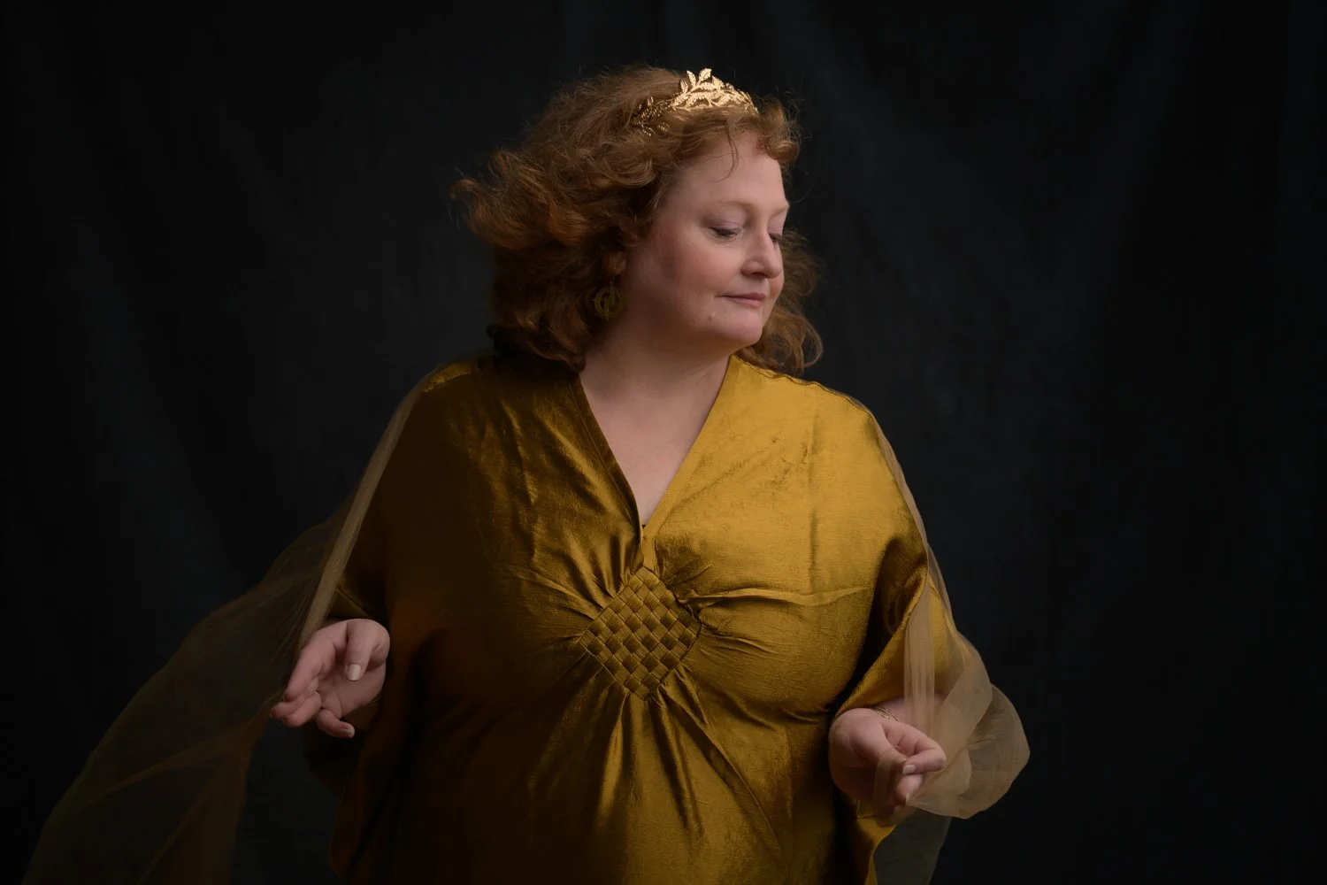 Woman in a flowing gold dress and crown posing against a black backdrop during a Goddess portrait session