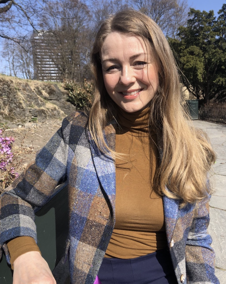 Ung kvinne sitter på en benk utendørs under solskinn, smiler og har på seg en sjakett og brunt turtleneck.