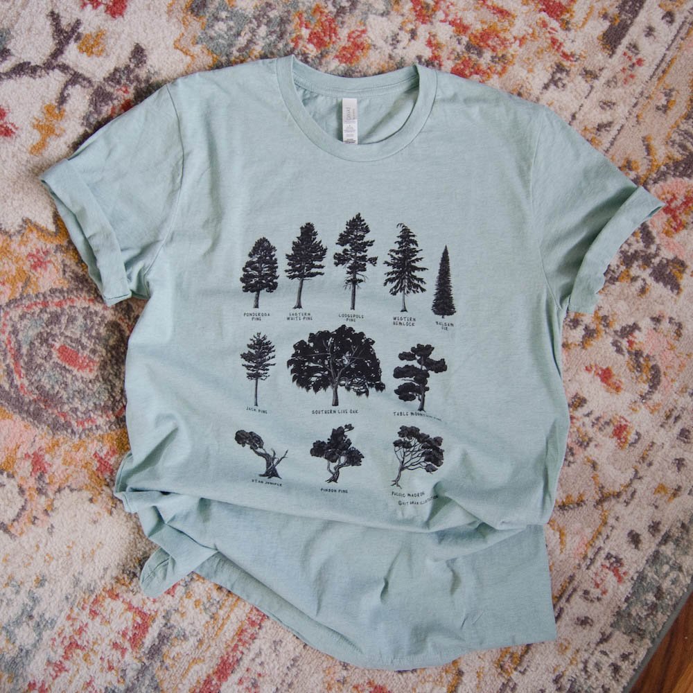 Pale dusty blue short-sleeve tee with black tree silhouettes is lying on a rug. Full design is visible, sleeves rolled up.