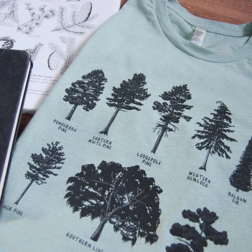 Close up of a folded pale dusty blue tee with black tree silhouette drawings. Field guide is in the background.