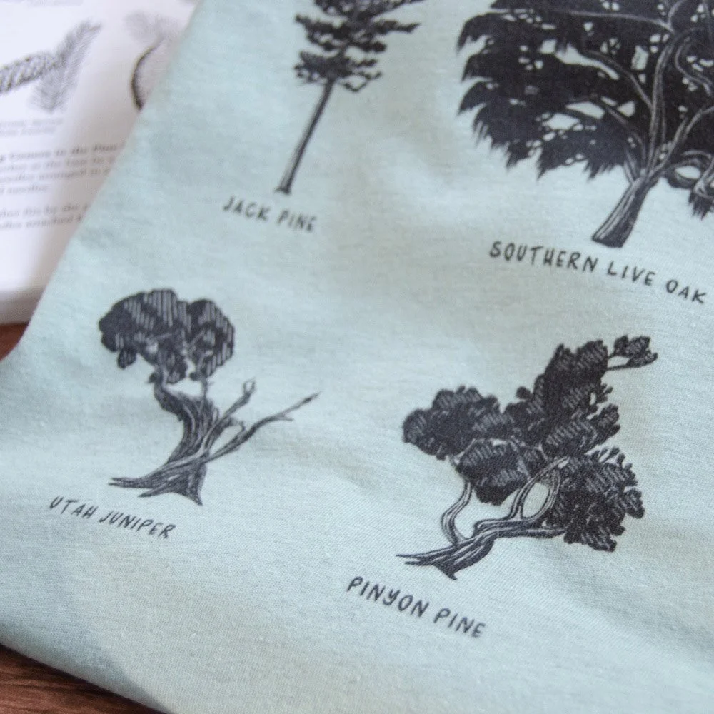 Close up of a folded pale dusty blue tee with black tree silhouette drawings. Utah juniper and pinyon pine are seen on tee.