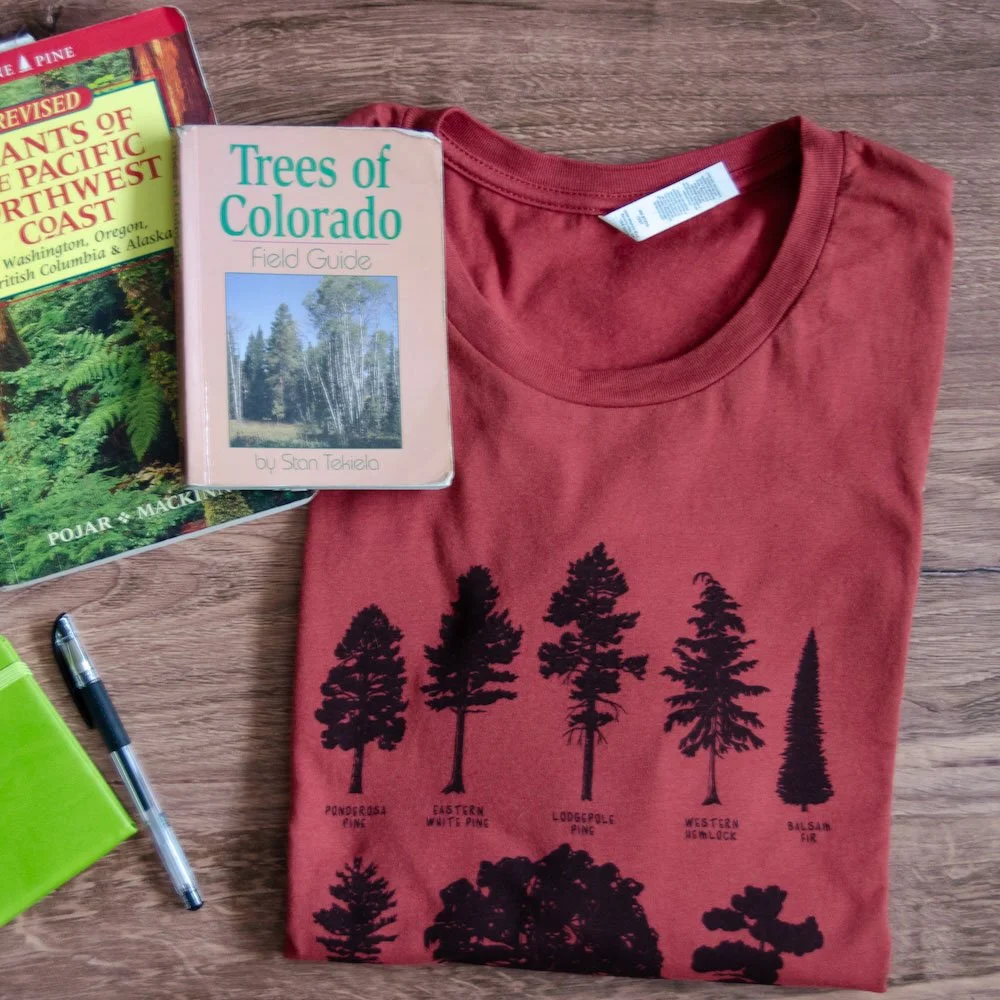 Folded rust red tee with black evergreen tree silhouette drawings sits next to field guides, a notebook, and pen.