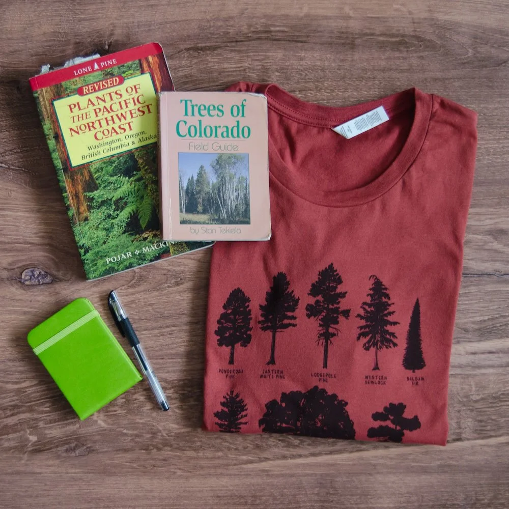 Folded rust red tee with black evergreen tree silhouette drawings sits next to field guides, a notebook, and pen.