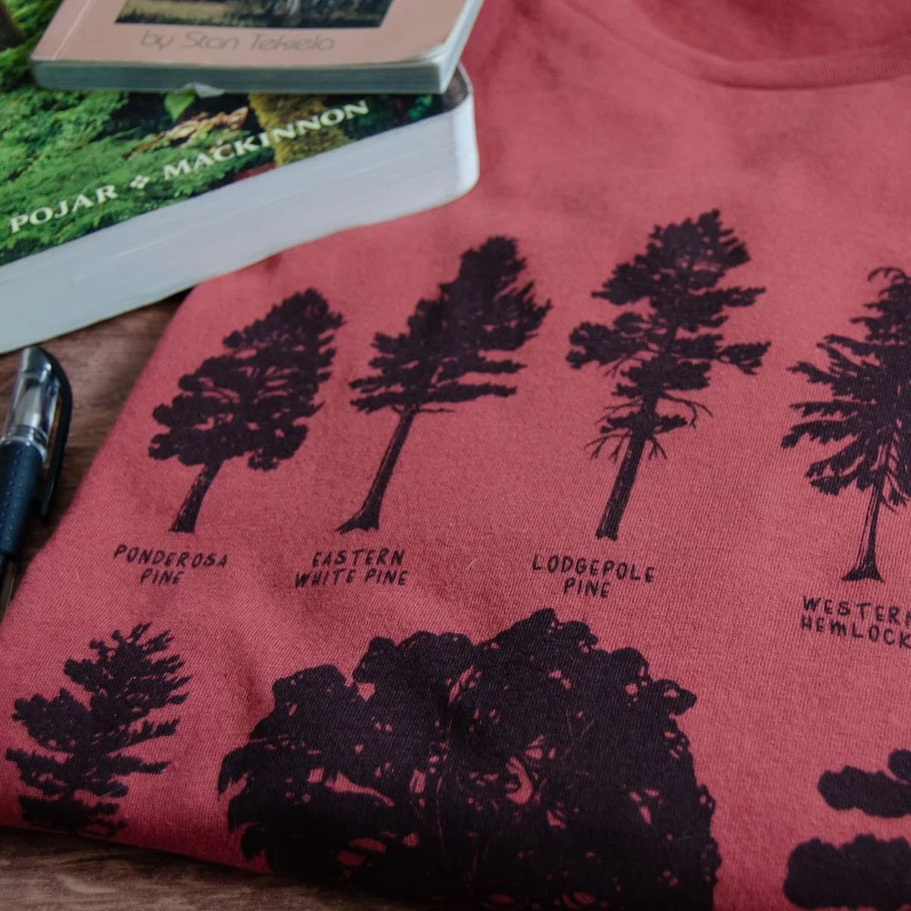 Close up of a folded rust red tee with black tree silhouette drawings. Field guides are in the background.