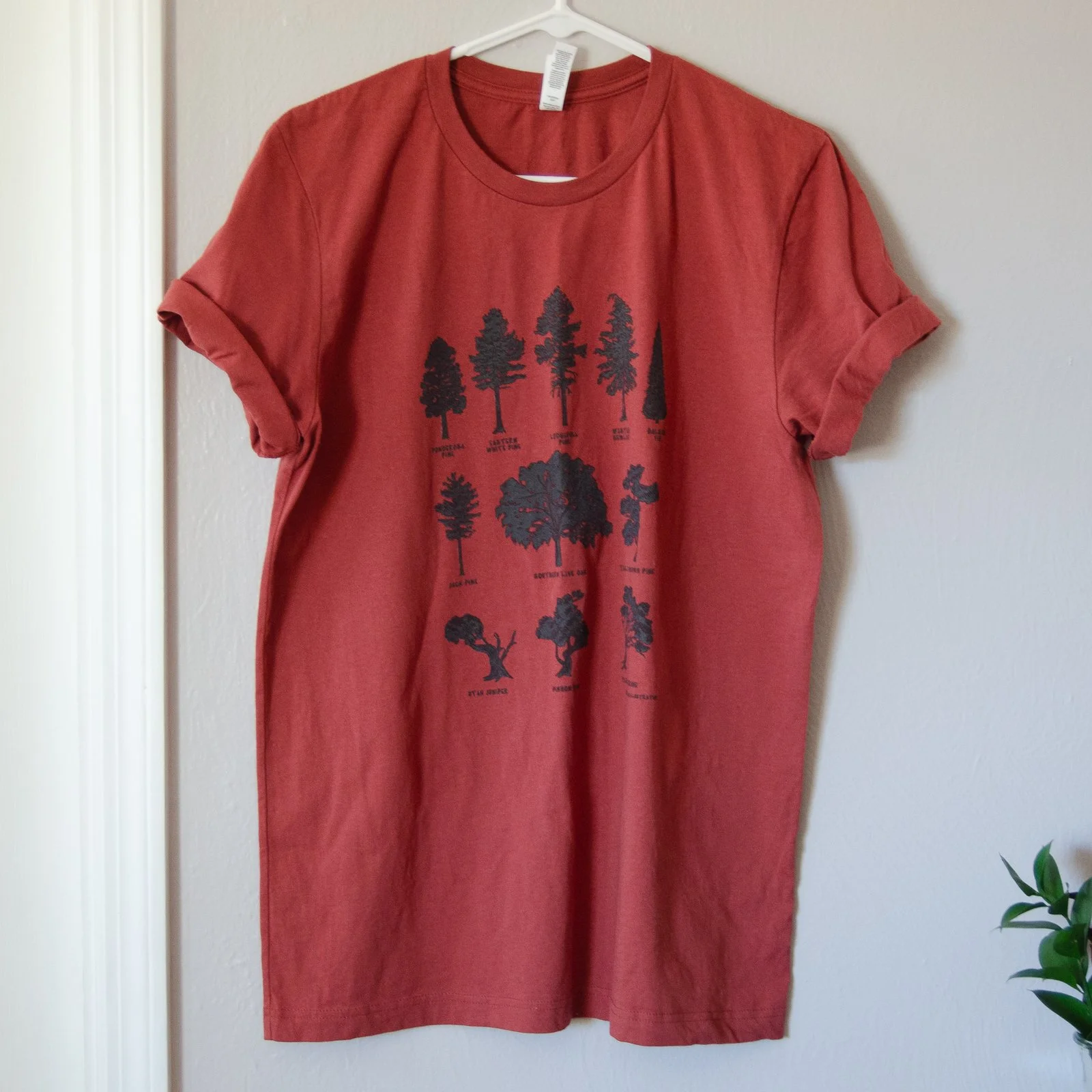 Rust red short-sleeve tee with 11 black tree silhouettes is hanging up on a hanger in front of a wall, showing entire design.