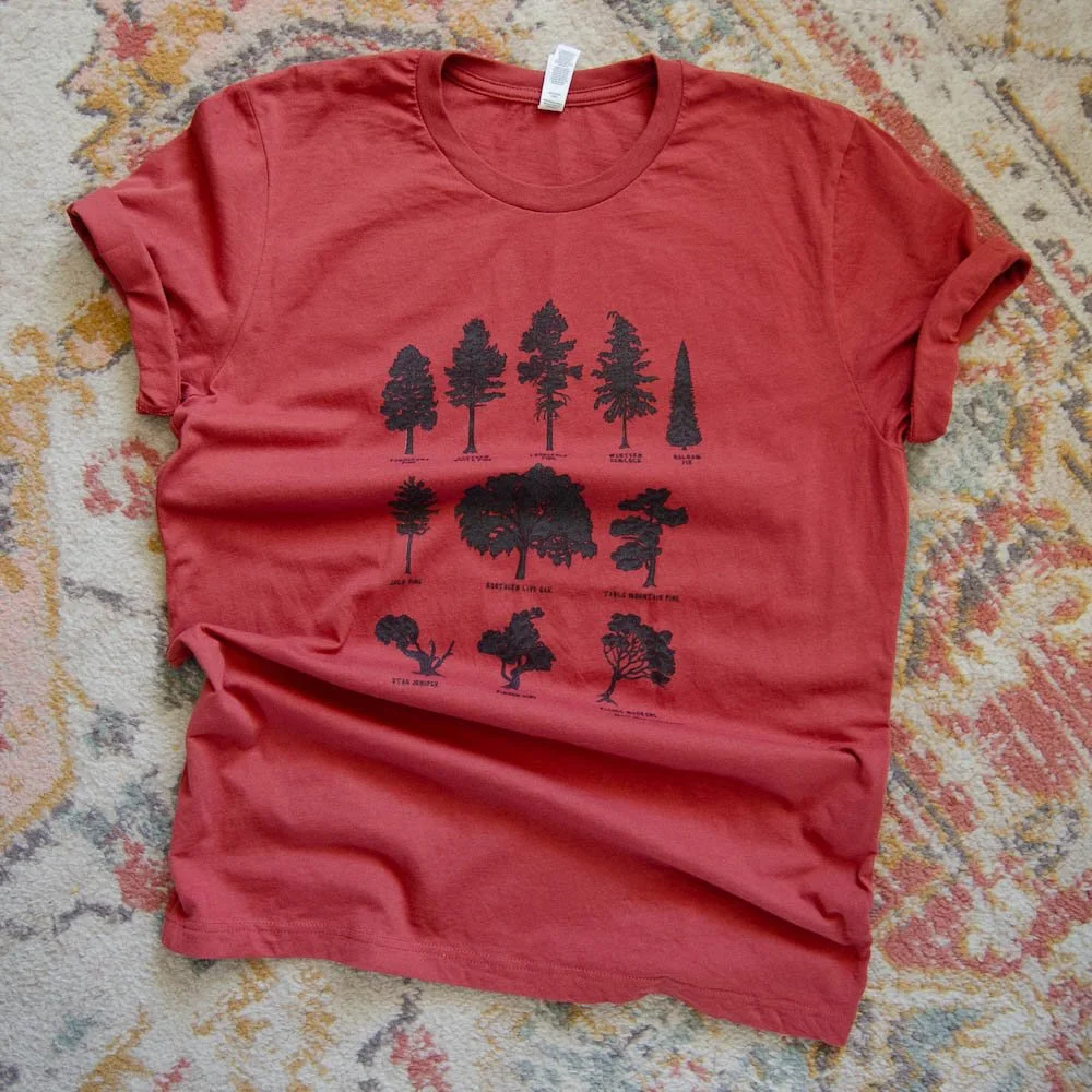 Rust red short-sleeve tee with 11 black tree silhouettes is lying on a rug. Full design is visible, sleeves rolled up.