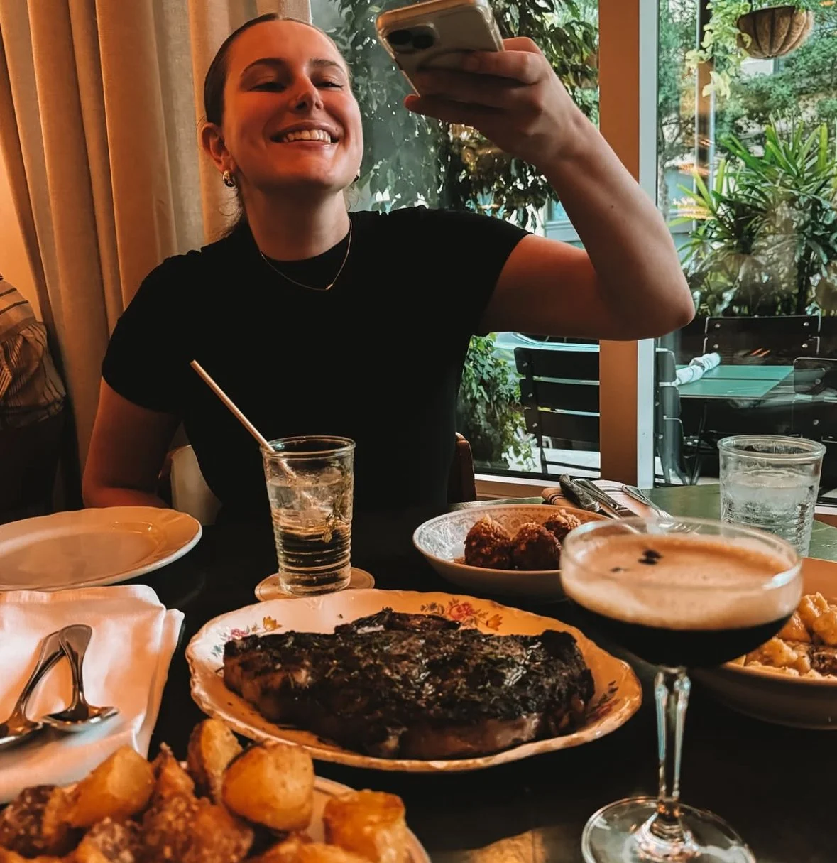 Taylor Oliver of Tayrex Creative (based in Baton Rouge, Louisiana) is seen taking a photo of the food on a table at a restaurant. She is wearing a black dress, gold jewelry, and her hair in a slicked back pony.