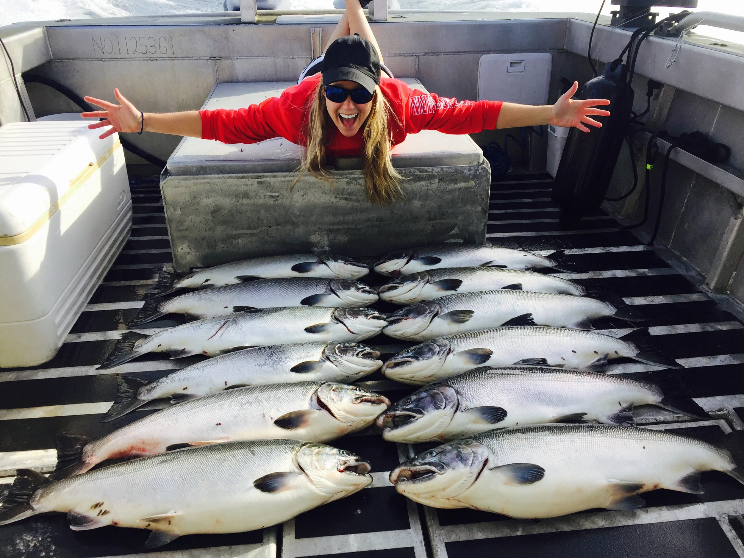 A woman on a boat enthusiastically shows off her catch of large fish laid out on the boat floor, with her arms wide open, wearing sunglasses, a black cap, and a red long sleeve shirt.