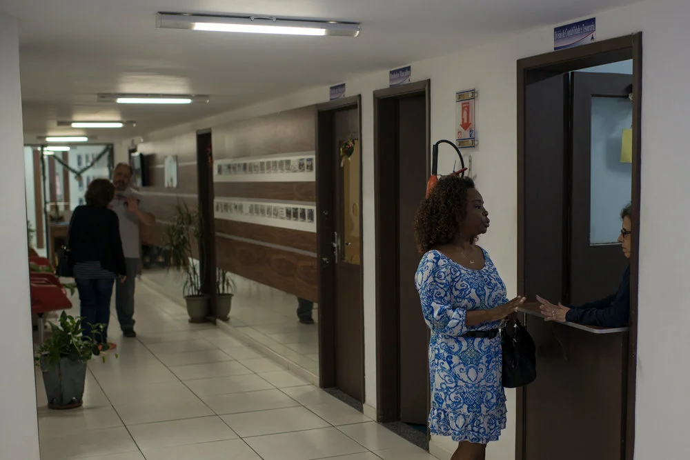 Dez. 6, 2017. Na mesma manhã, Sarah conversa com funcionária da tesouraria na sede da FEVRE (Fundação Educacional de Volta Redonda), que é responsável pela Ensino Fundamental e Médio da cidade. As funções como acompanhar os trâmites políticos relacionados às verbas e checar liberação de veículos para transporte dos alunos e dos instrumentos em dias de concerto ficam por conta da Sarah, que tem boa abertura nos segmentos administrativos municipais por conta dos mais de 20 anos em que atua no projeto.
