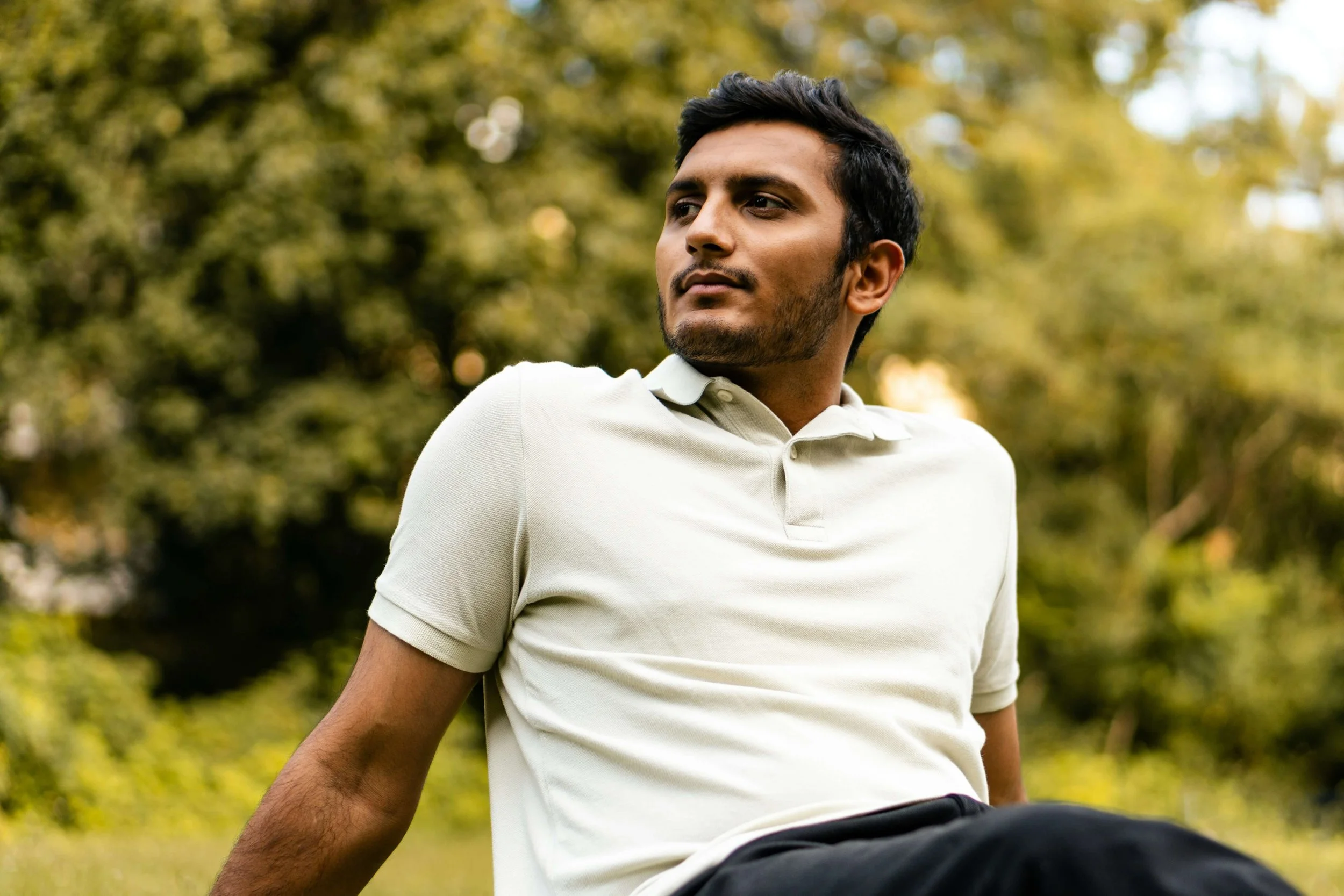 Man sitting outdoors on grass with trees in background, wearing a light colored polo shirt.