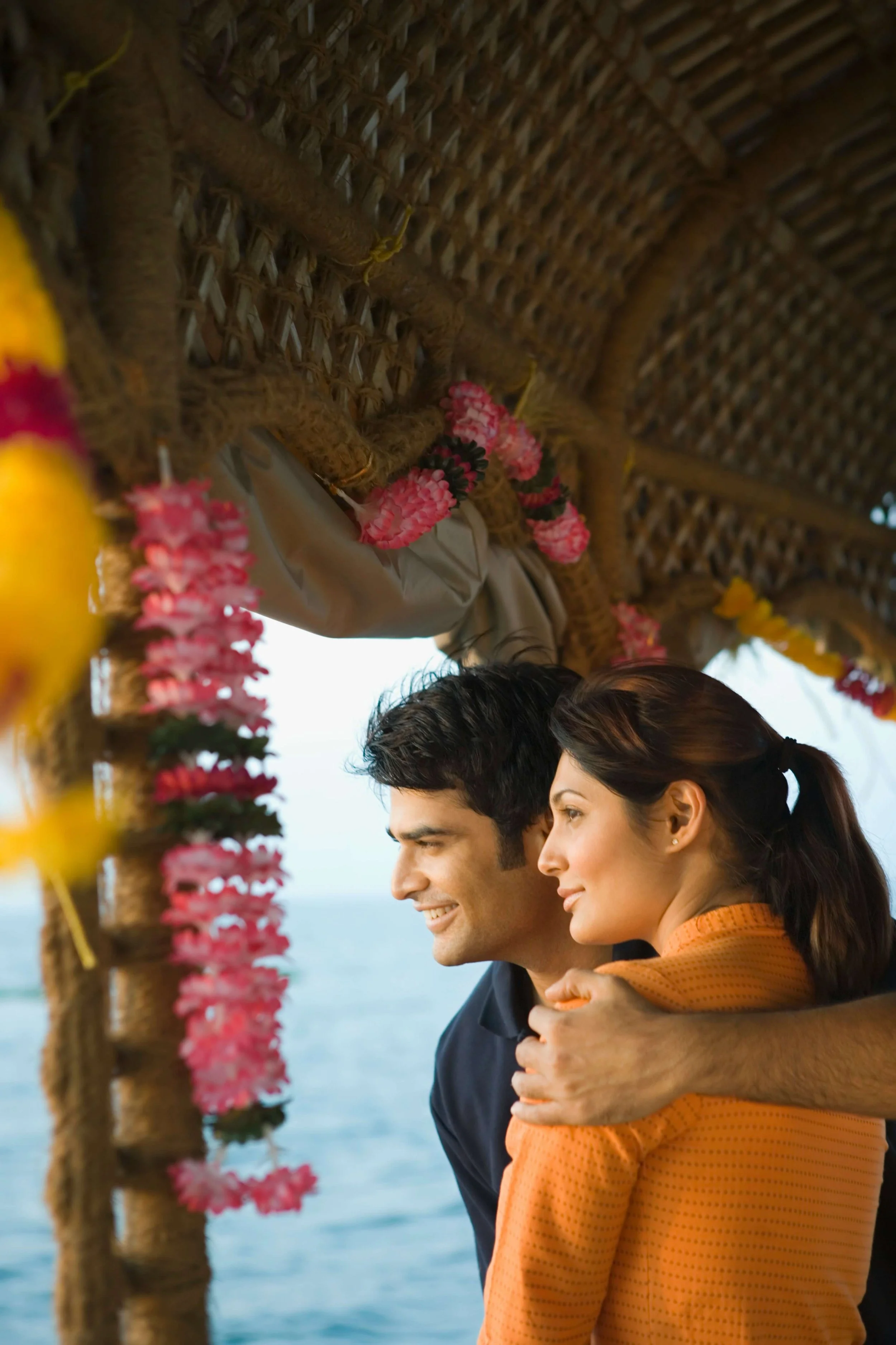 A young couple embracing on a pier decorated with pink flowers, with the ocean and sky in the background.