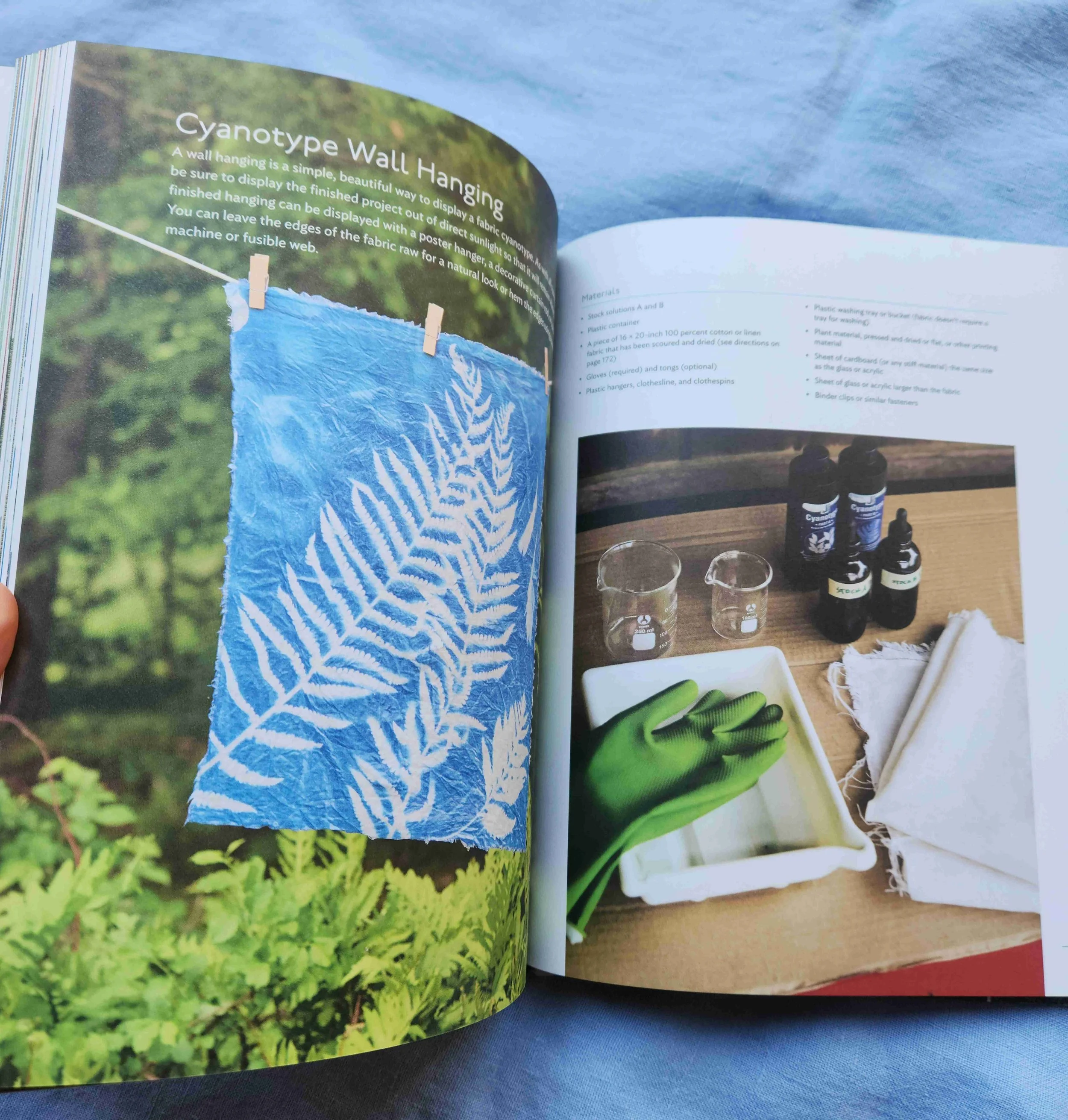 Open book displaying instructions for cyanotype wall hanging, including materials like fabric, chemicals, gloves, and tools, with a photo of the finished cyanotype hanging on a line and a workspace with chemicals and supplies.
