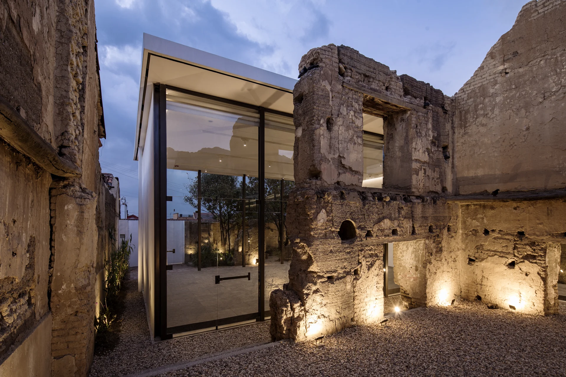 La forma del piso y el espacio resultaron de ocupar el negativo de lo que antiguamente fuera la casa, invirtiendo el adentro y el afuera, encapsulando las ruinas como si fueran reliquias. En resonancia respetuosa con la obra de arte, las fachadas se diseñaron con el máximo formato local de vidrio y una única esbelta estructura metálica que le da soporte a la ventanearía y la cubierta. El piso compuesto de 1296 tabletas de 60cm x 60cm, hechas con el metal fundido de las armas, se pliega en los bordes, creando una superficie monolítica de 16 cm de altura. La piel del espacio, modulada a distancia de 2.40 m se dilata hacia el interior en los laterales para dejar ver la robustez de la pieza y desaparecer frente a la ruina. Hacia afuera, aterriza sobre una losa de concreto esmaltado a ras de los patios en piedra.