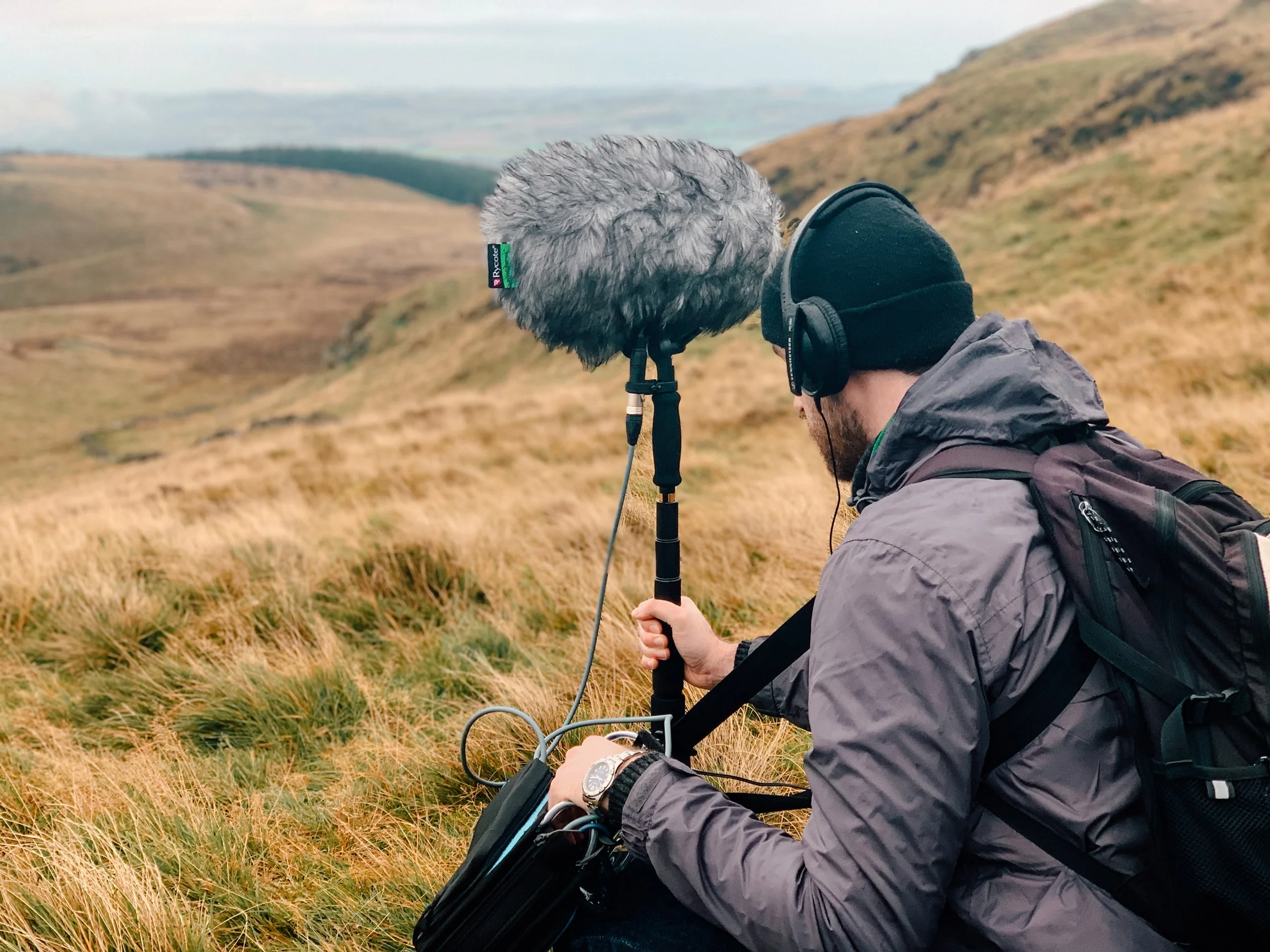 A man wearing a black beanie, gray jacket, and backpack is sitting in a grassy field with rolling hills in the background. He is holding a portable audio recording device with a large windscreen microphone attached.