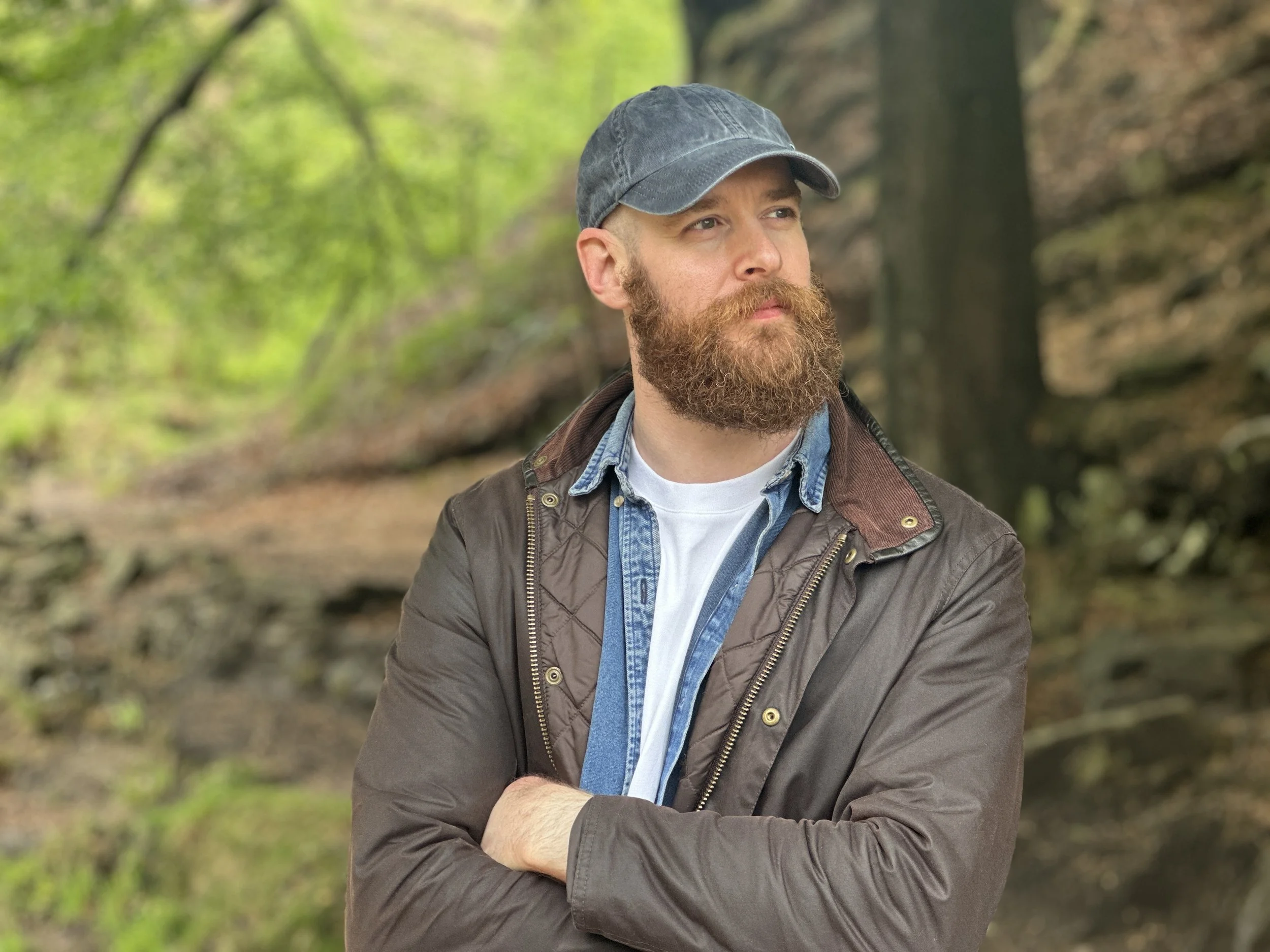 Man with brown beard wearing a denim cap, brown jacket, and layered shirts stands outdoors in a wooded area with trees and greenery.