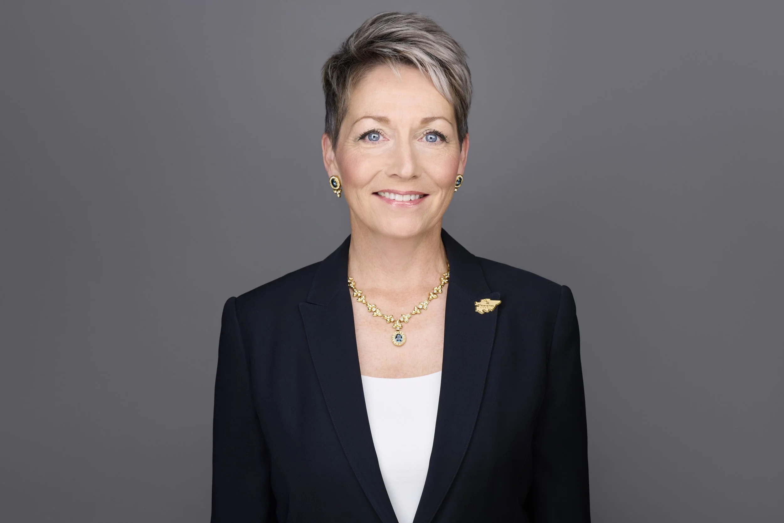 Woman with short grey and blonde pixie-cut hair in a black blazer with a gold Sturgeon County pin, wearing a gold and blue gemstone necklace and earrings, corporate headshot against a grey background.