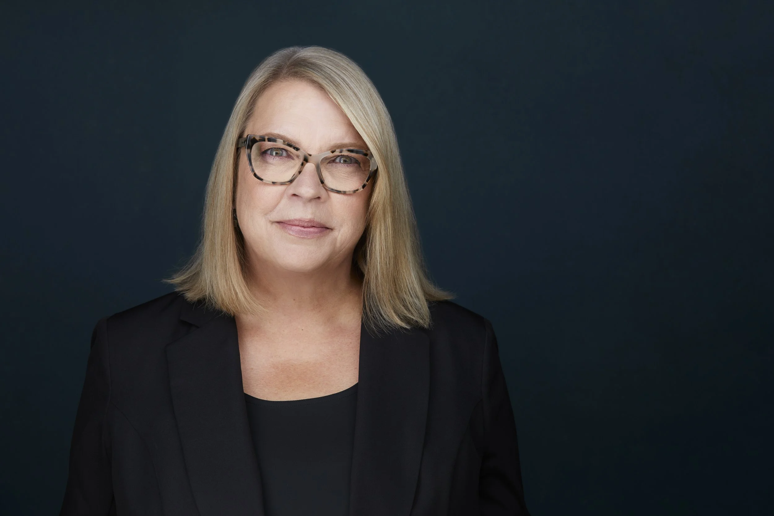 Woman with blonde shoulder-length hair and tortoiseshell glasses in a black blazer, corporate headshot against a dark blue background.