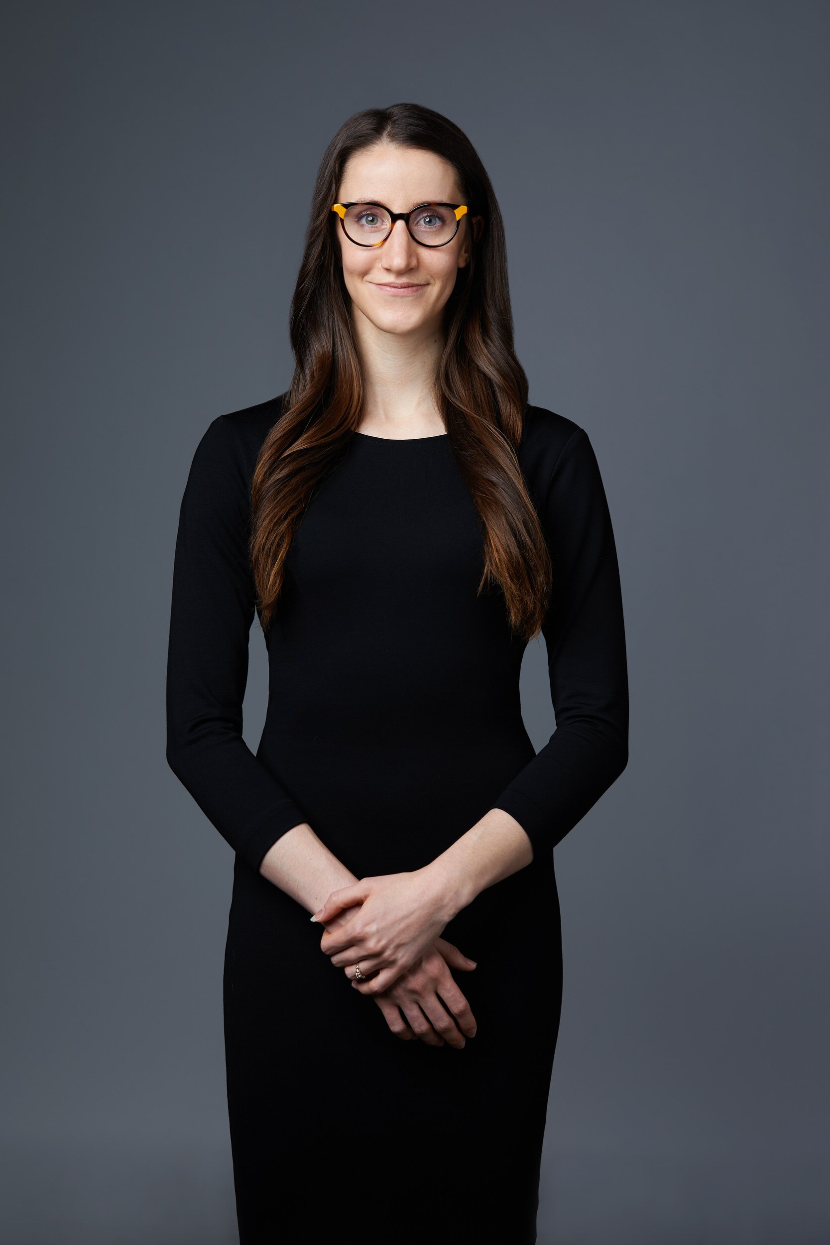 Woman with long dark hair and round glasses in a black dress, hands clasped, standing business portrait against a grey background