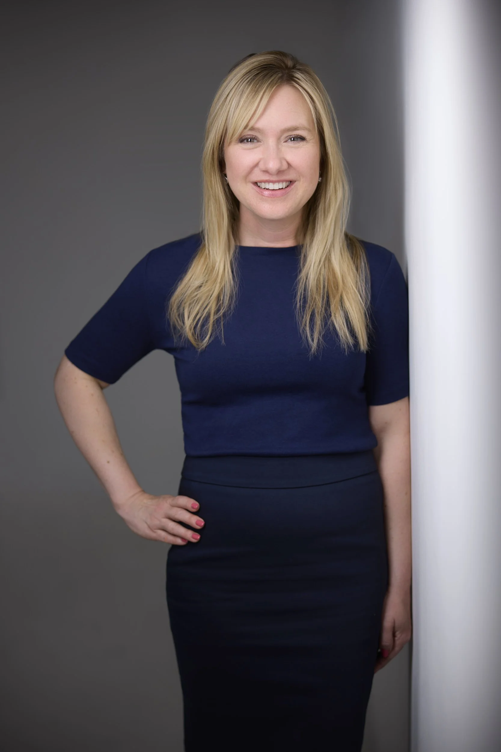Woman with long blonde hair, smiling, standing with a hand on her hip in a navy top and skirt, business portrait against a grey background.