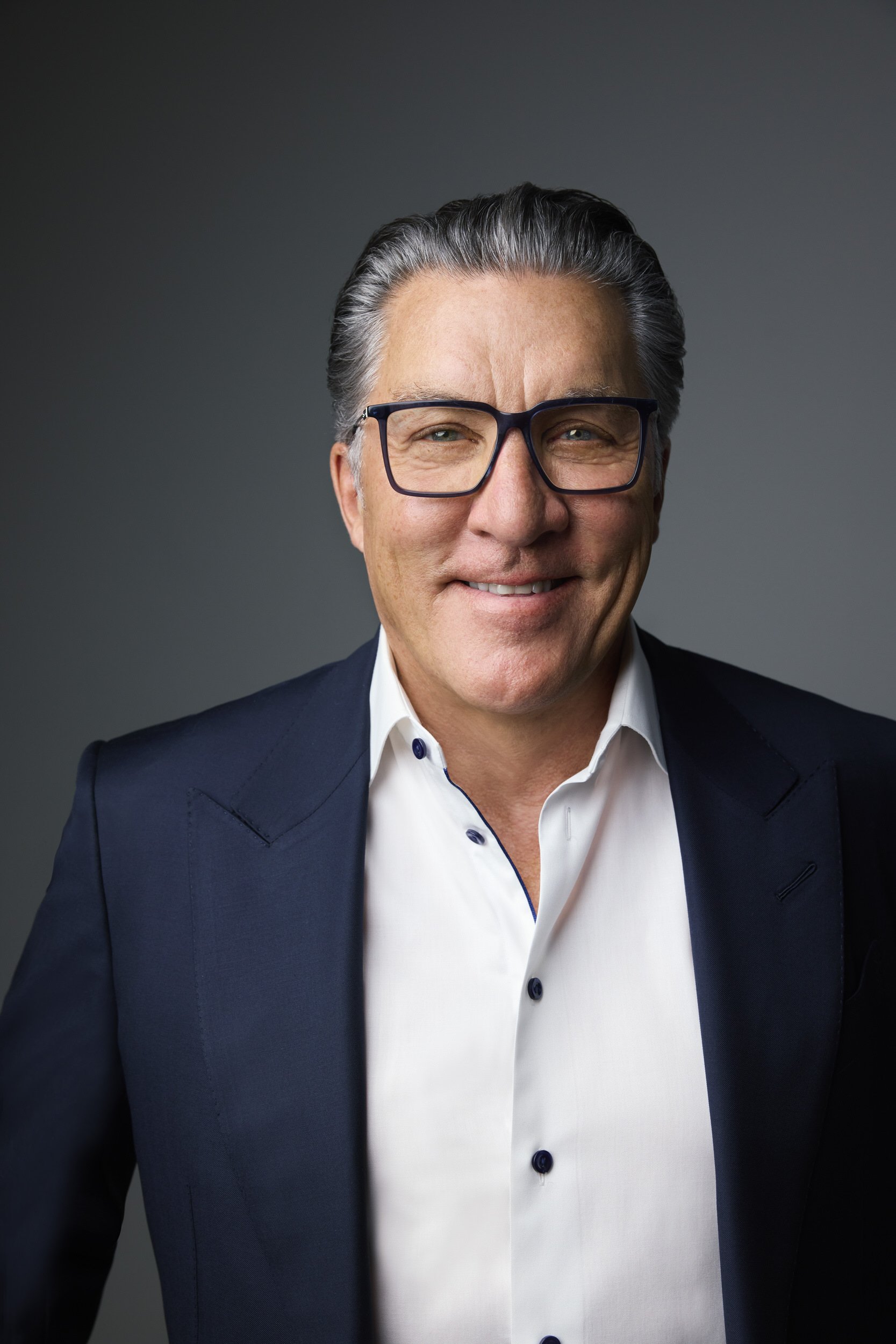 Man with grey hair and dark-framed glasses in a navy blazer and white shirt, smiling corporate headshot against a grey background.