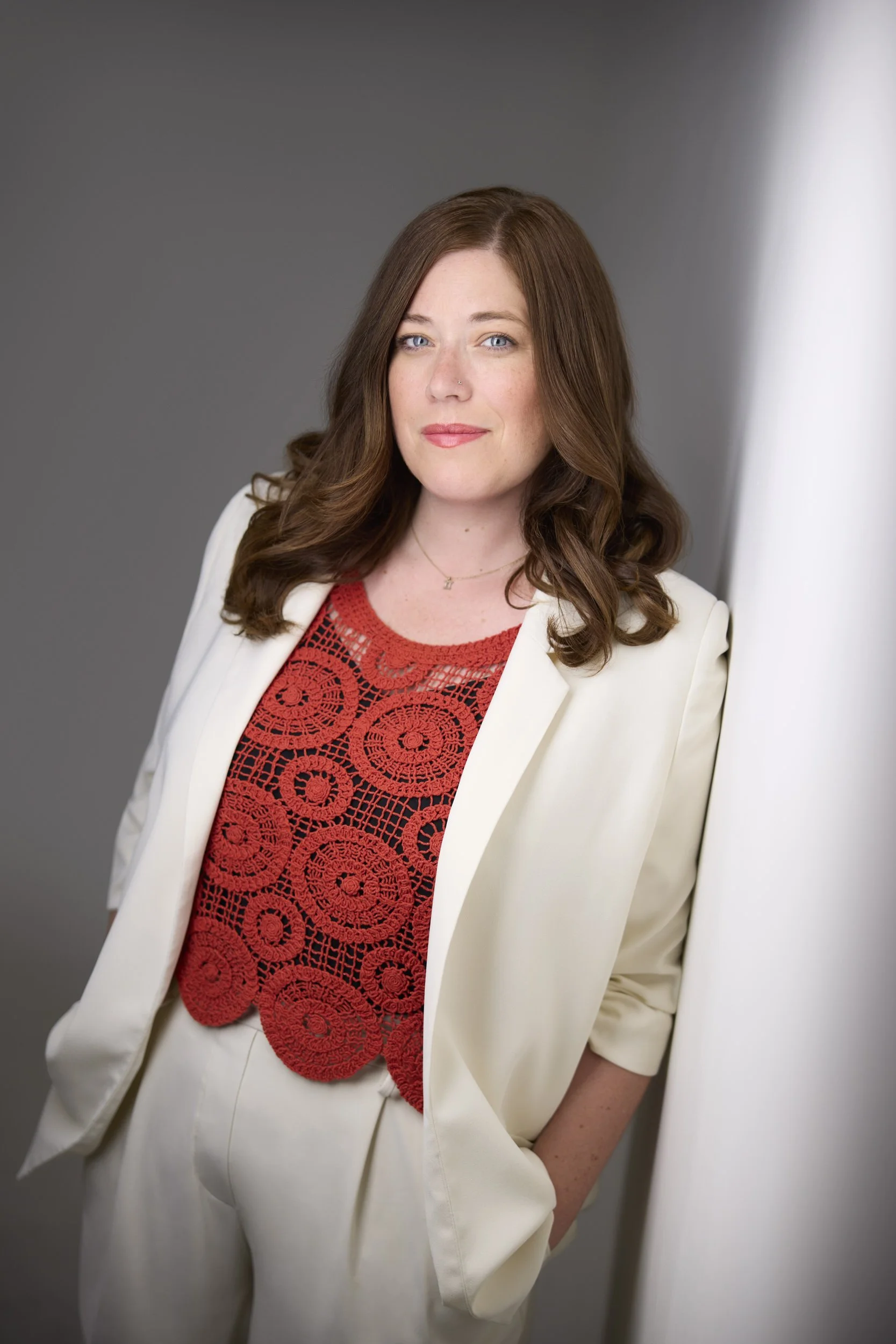 Woman with long brown hair in a cream blazer and red crochet top, business portrait against a grey background.