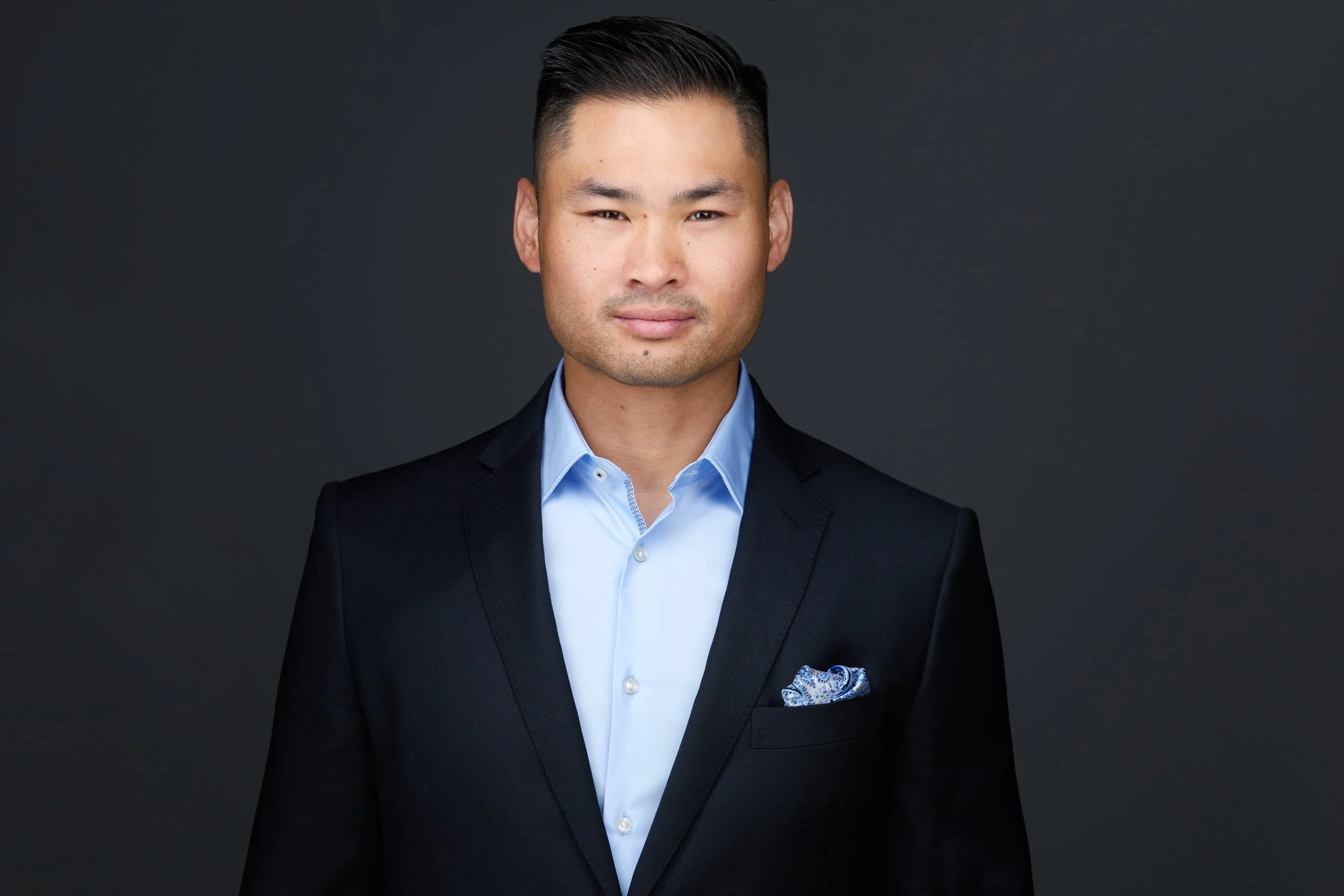 Man with short dark hair in a black suit with a light blue shirt and patterned pocket square, corporate headshot against a dark grey background.