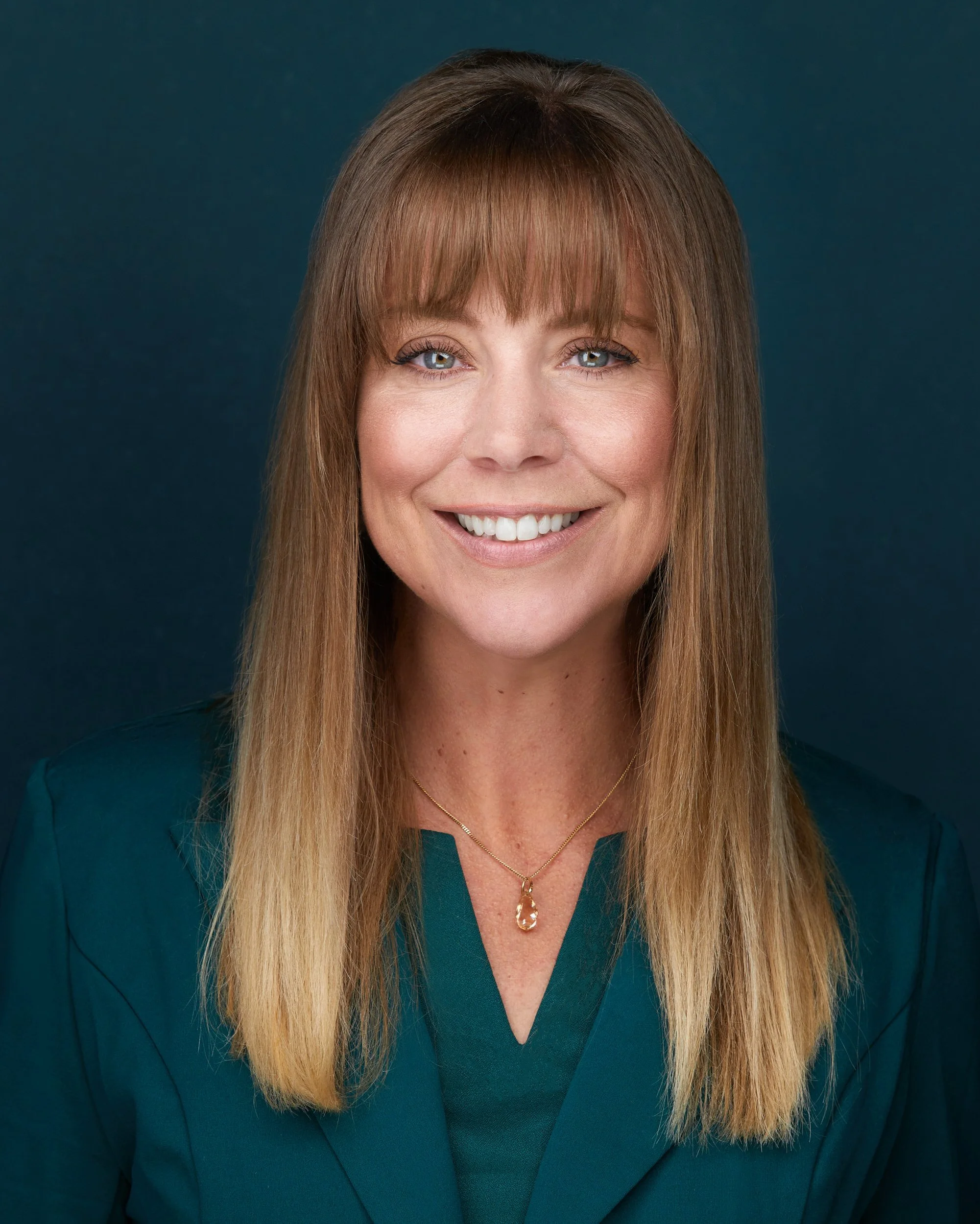 Woman with long light brown hair and bangs in a teal blazer with a gold necklace, smiling headshot against a dark teal background