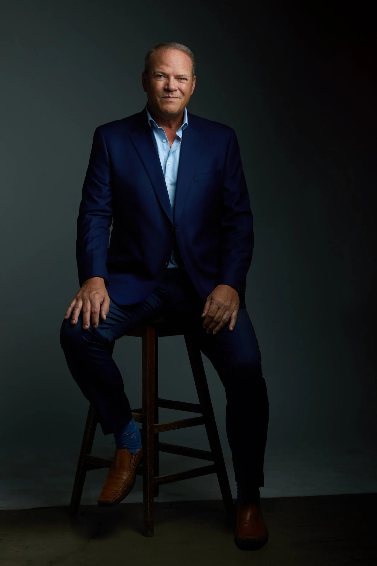 Man in a navy suit and light blue shirt, seated full-body business portrait against a dark background