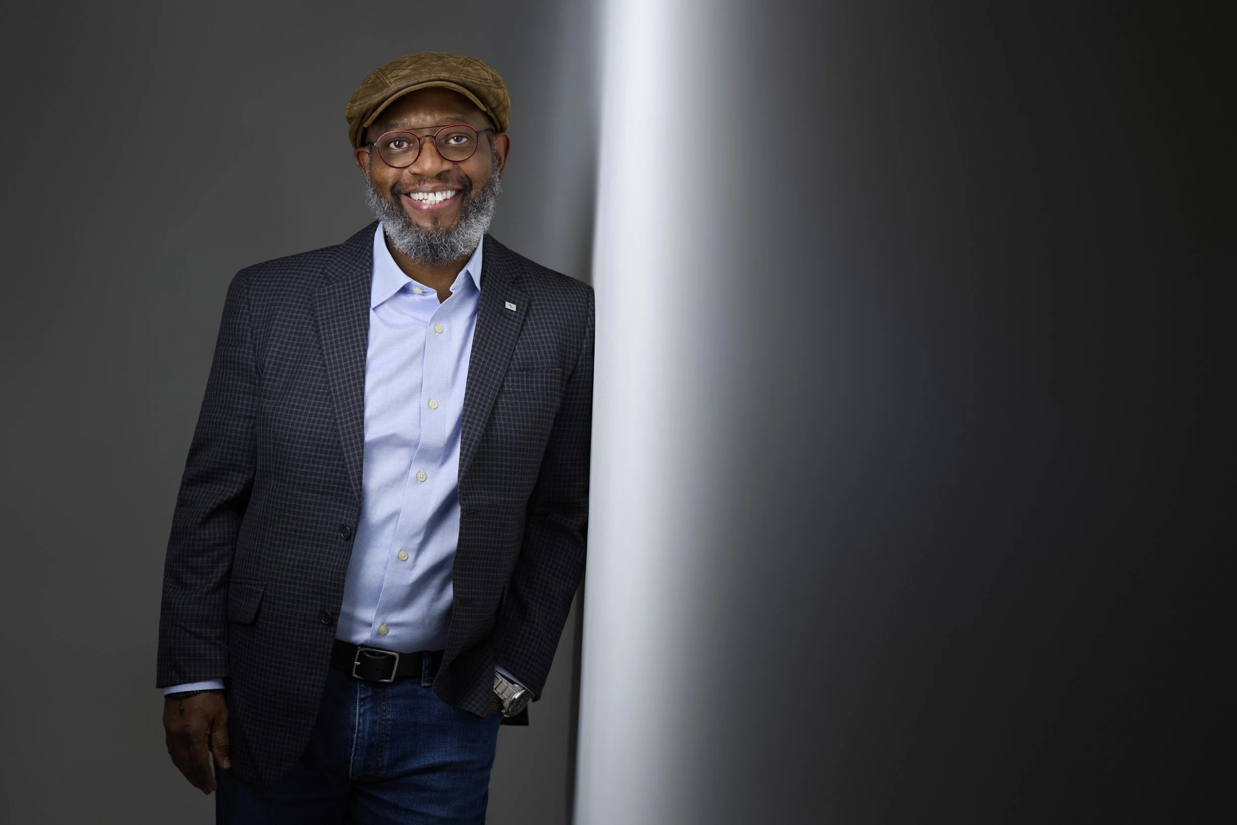 Man with a beard wearing glasses and a flat cap, navy checkered blazer, smiling headshot against a grey background.